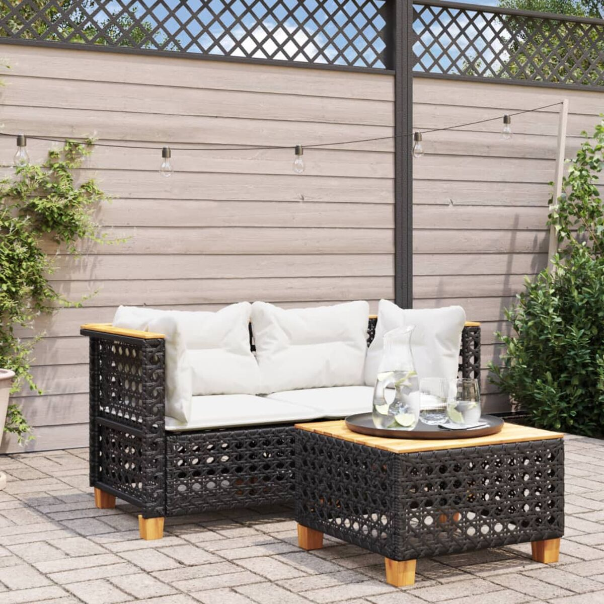 Schwarzes Outdoor-Sofaset aus Korbgeflecht mit weißen Kissen und einem Holztisch. Gepflasterte Terrasse.