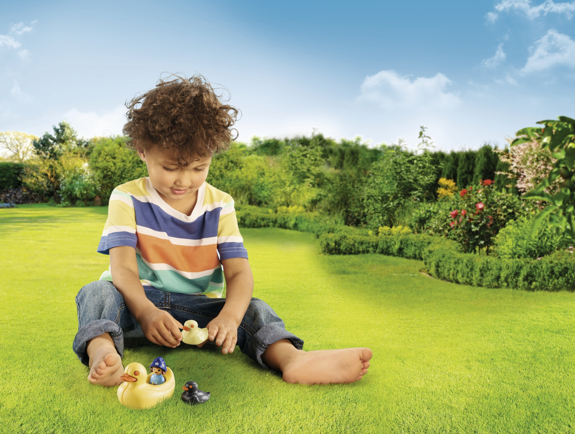 Ein kleiner Junge spielt mit Spielzeugenten auf einer grünen Wiese mit blauem Himmel im Hintergrund.