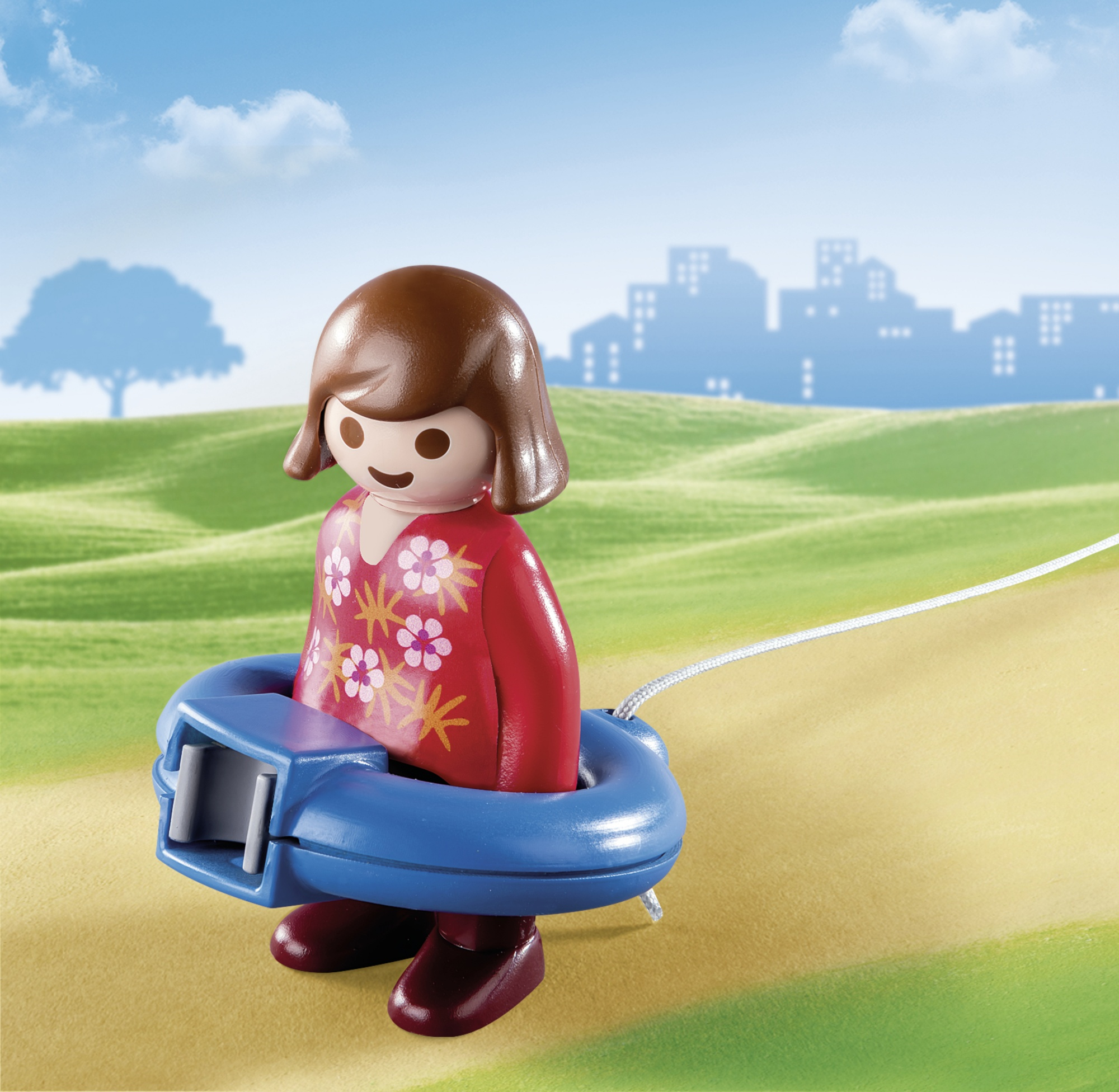 Eine Playmobil-Mädchenfigur geht auf einem Pfad und hält einen blauen, runden Drachenaufwickler mit Schnur. Stadt im Hintergrund.
