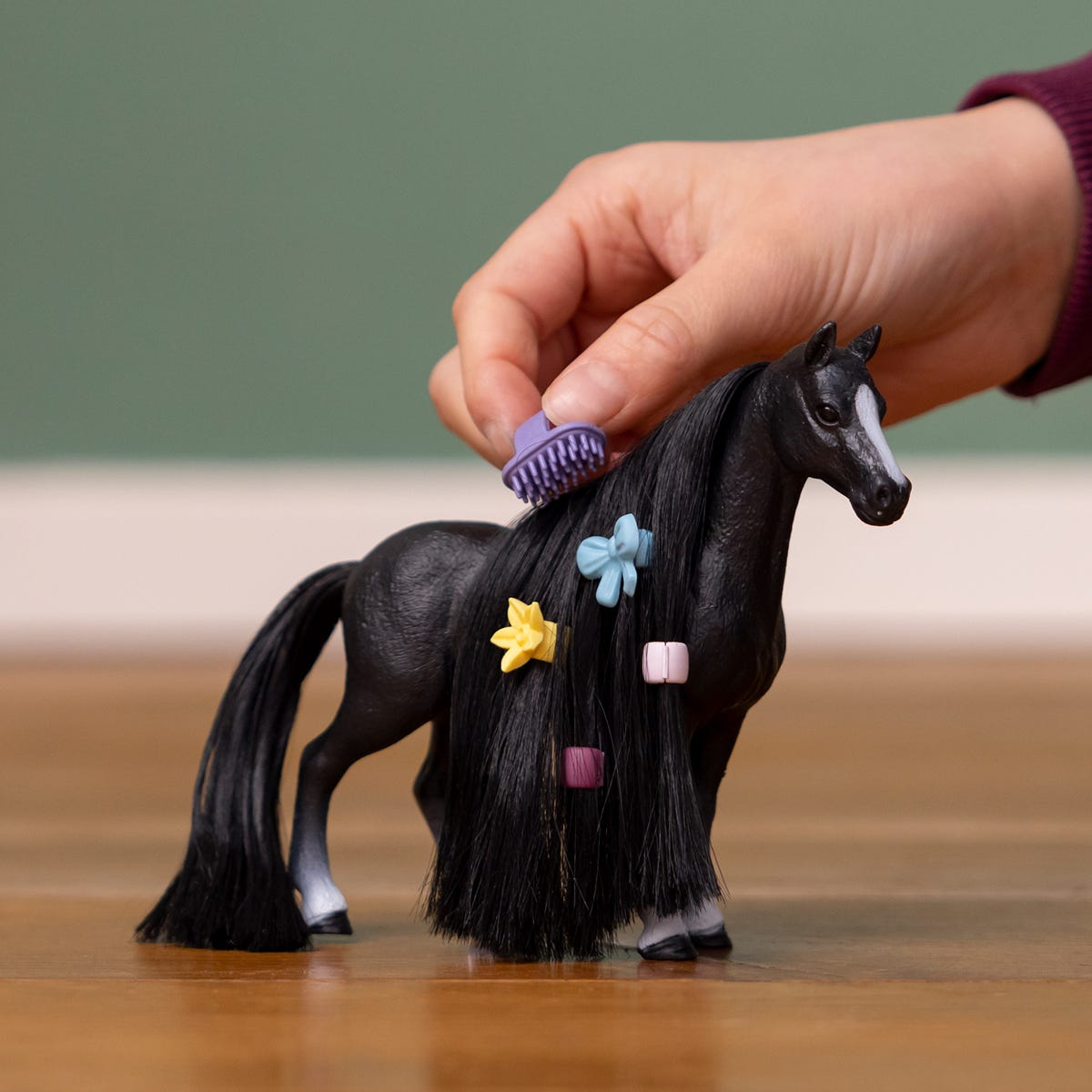 Die Hand einer Person bürstet die Mähne eines schwarzen Spielzeugpferdes. Auf der Mähne des Pferdes befinden sich bunte Haaraccessoires.