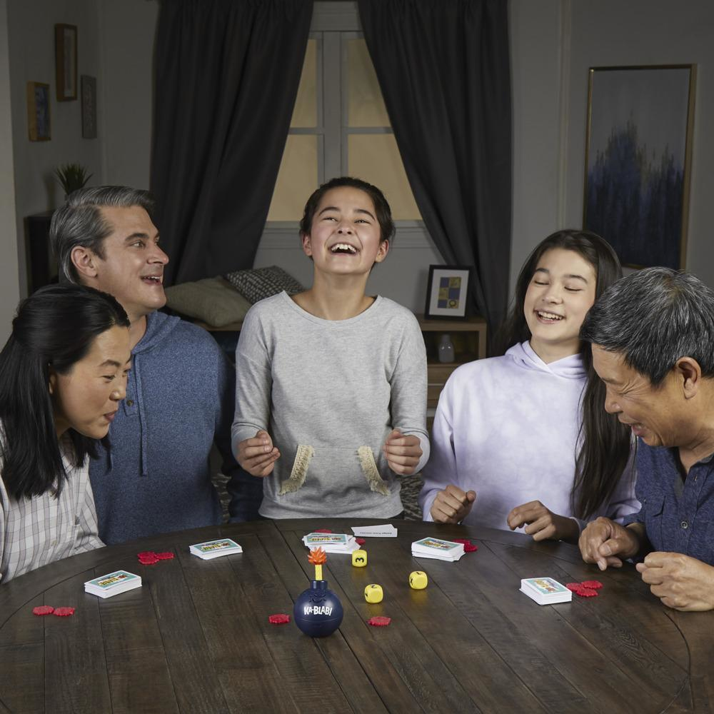 Fünf Leute lachen um einen Tisch herum und spielen ein Spiel. Karten, Würfel und Spielsteine liegen auf dem Tisch.