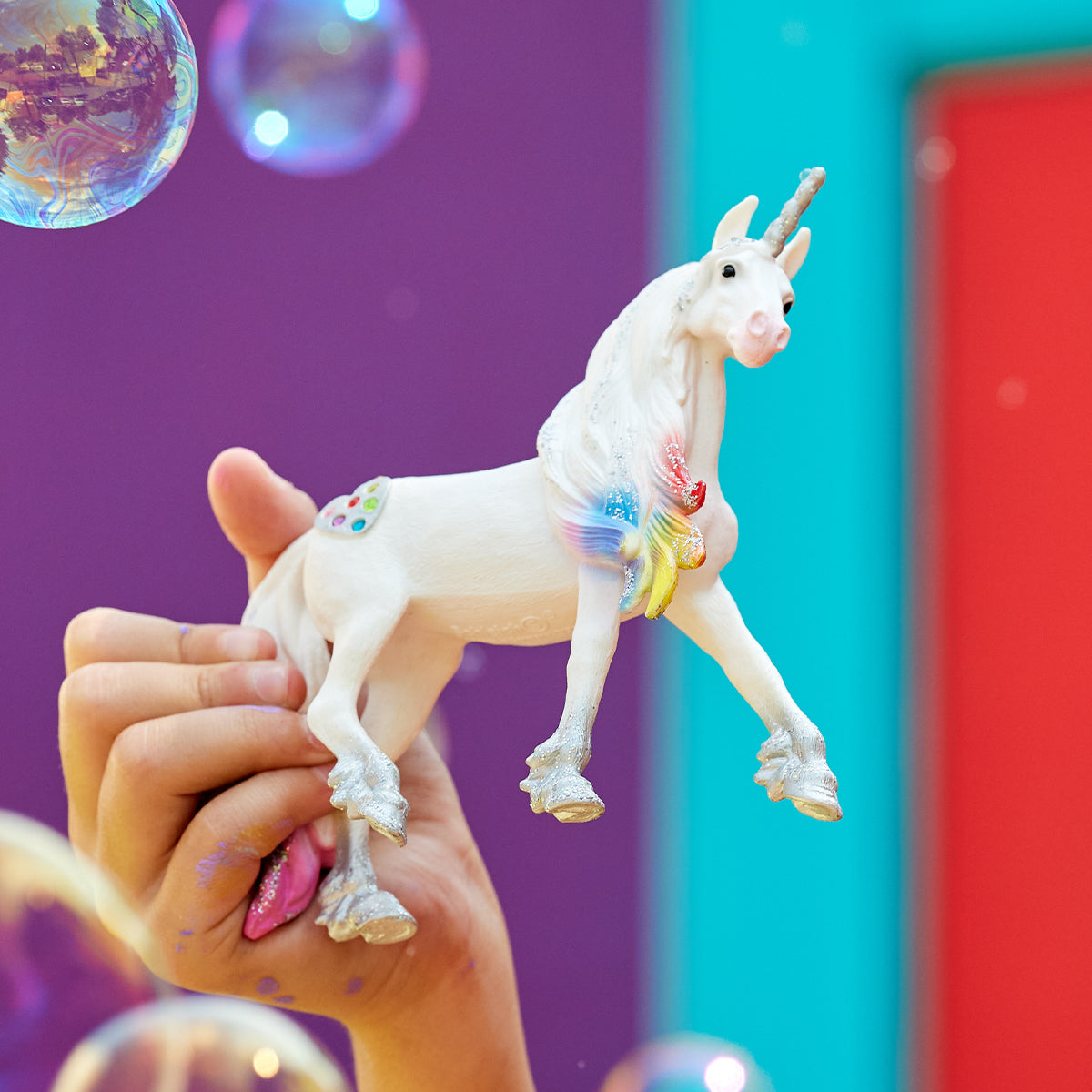 Ein weißes Spielzeug-Einhorn mit Regenbogenmähne und -schwanz, gehalten von einer Hand, mit Blasen im Hintergrund.