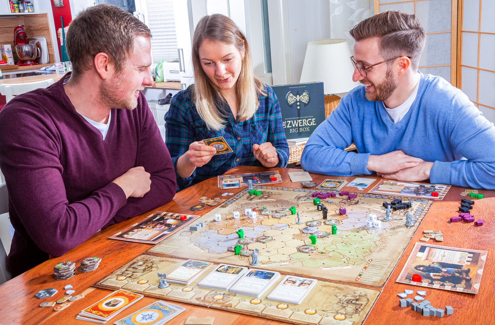 Drei Personen spielen ein Brettspiel an einem Holztisch, lächeln und interagieren.