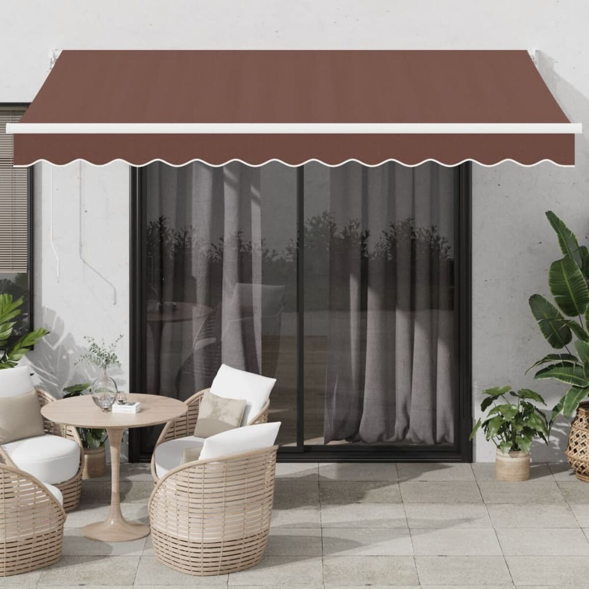 Una tenda da sole marrone sopra una porta-finestra con sedie bianche e un tavolo. Sono presenti piante verdi.