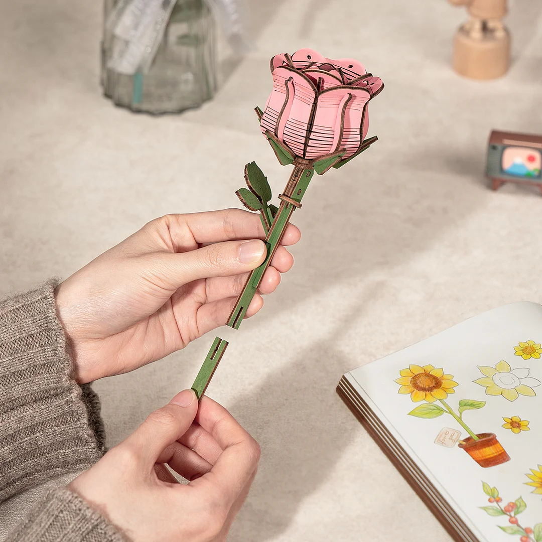 Handen die een houten rozenmodel in elkaar zetten met groene steel en roze bloemblaadjes.