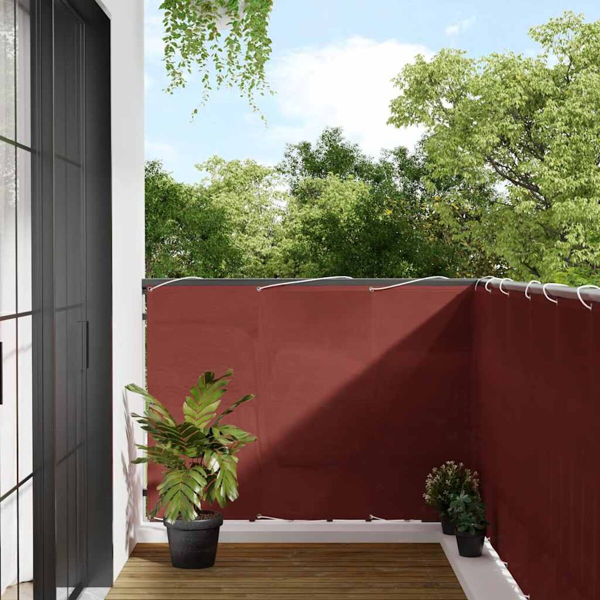 Un balcone con uno schermo per la privacy bordeaux, piante e uno scorcio di uno sfondo verde.