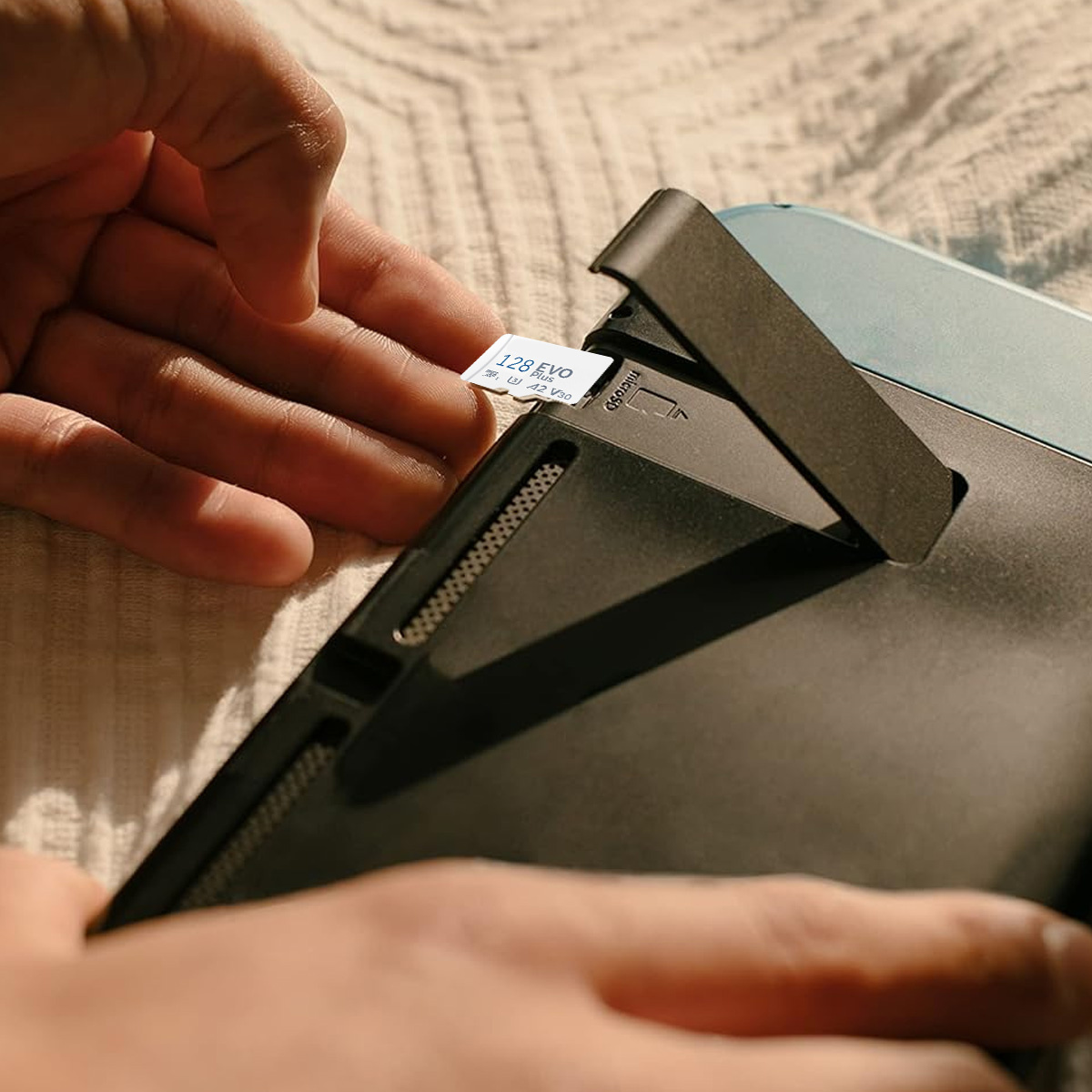 Eine Person, die eine 128GB Micro-SD-Karte in ein schwarzes Tablet einlegt. Das Tablet liegt auf einem Bett.