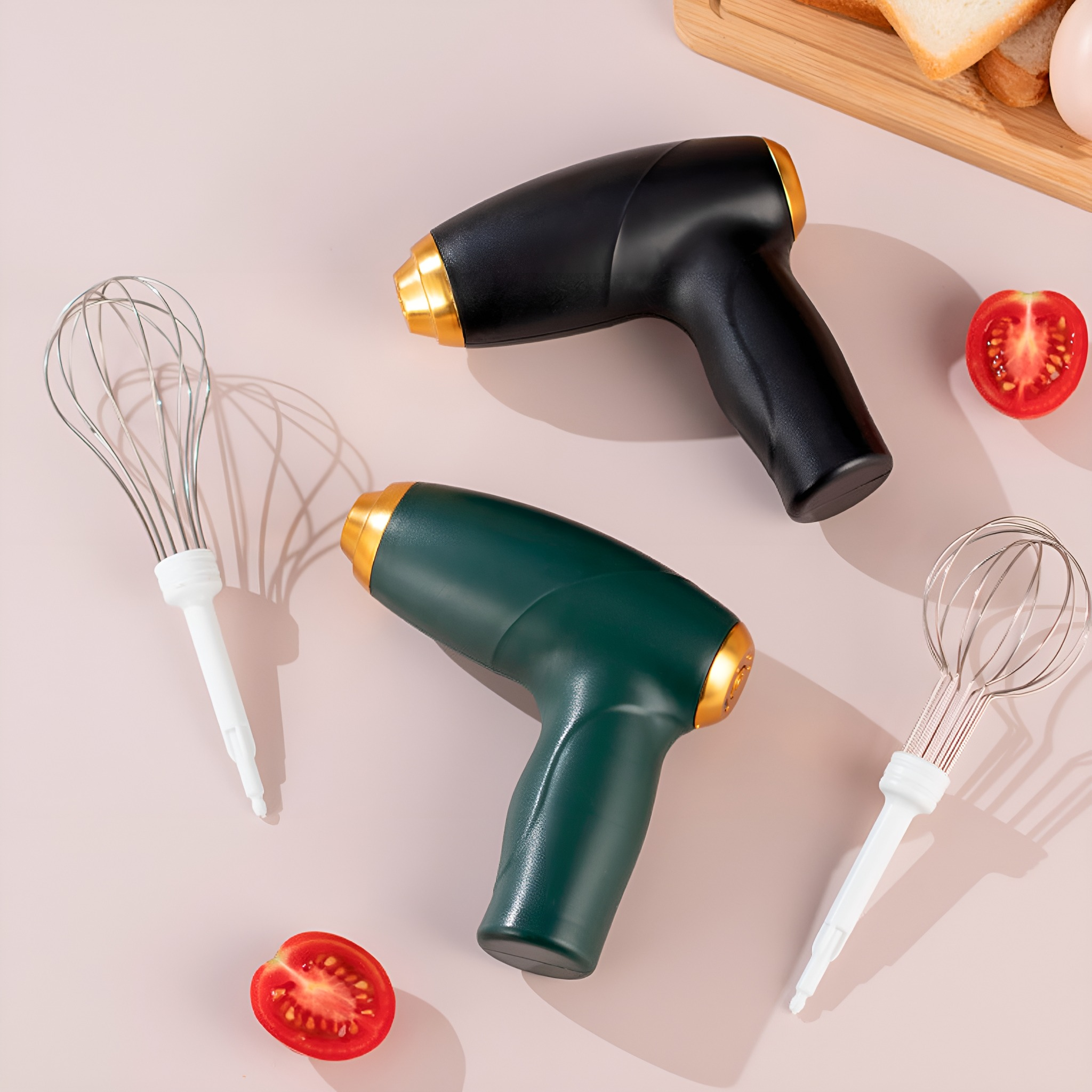 Zwei Handrührgeräte, eines schwarz und eines grün, mit goldenen Akzenten, sowie zwei Schneebesen und Tomaten.