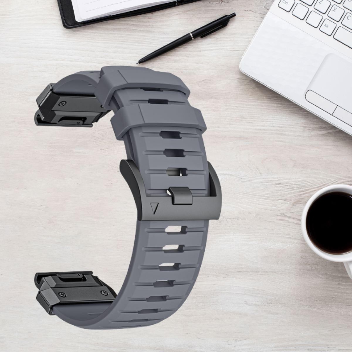Graues Uhrenarmband mit grauer Schnalle liegt auf einem hellen Holzschreibtisch mit Stift, Notizbuch und einer Tasse Kaffee.