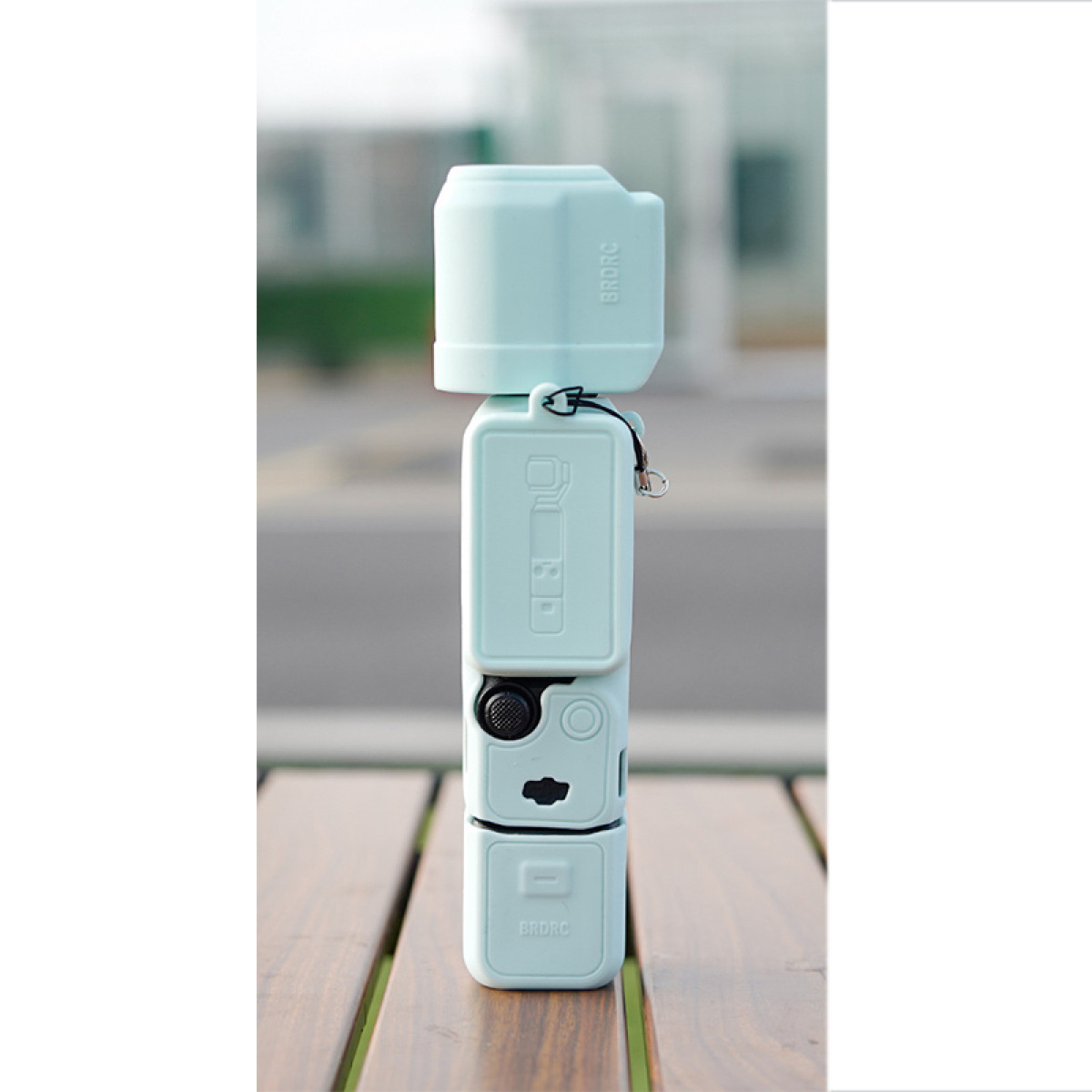 Un dispositivo azzurro simile a una fotocamera su una superficie di legno. Lo sfondo è sfocato, con alberi e un sentiero.