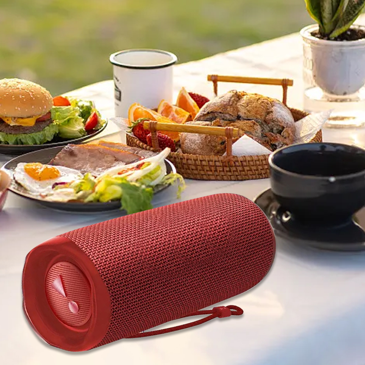 Roter tragbarer Lautsprecher auf einem Tisch mit Essen, darunter Burger, Salat, Brot und Orangen, im Freien.