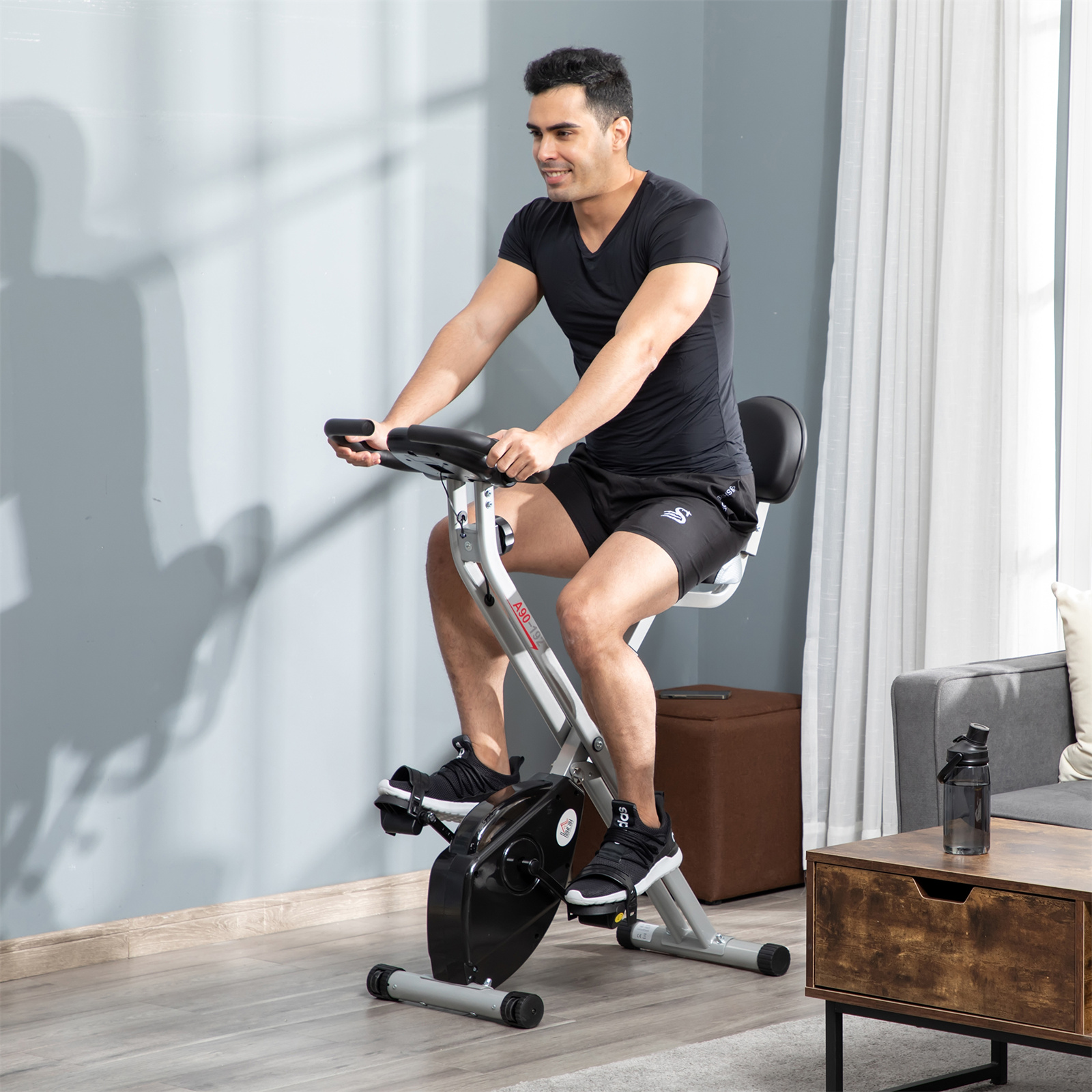 Ein Mann trainiert auf einem Heimtrainer. Er trägt ein schwarzes Hemd und Shorts. Auf einem Tisch steht eine Flasche.