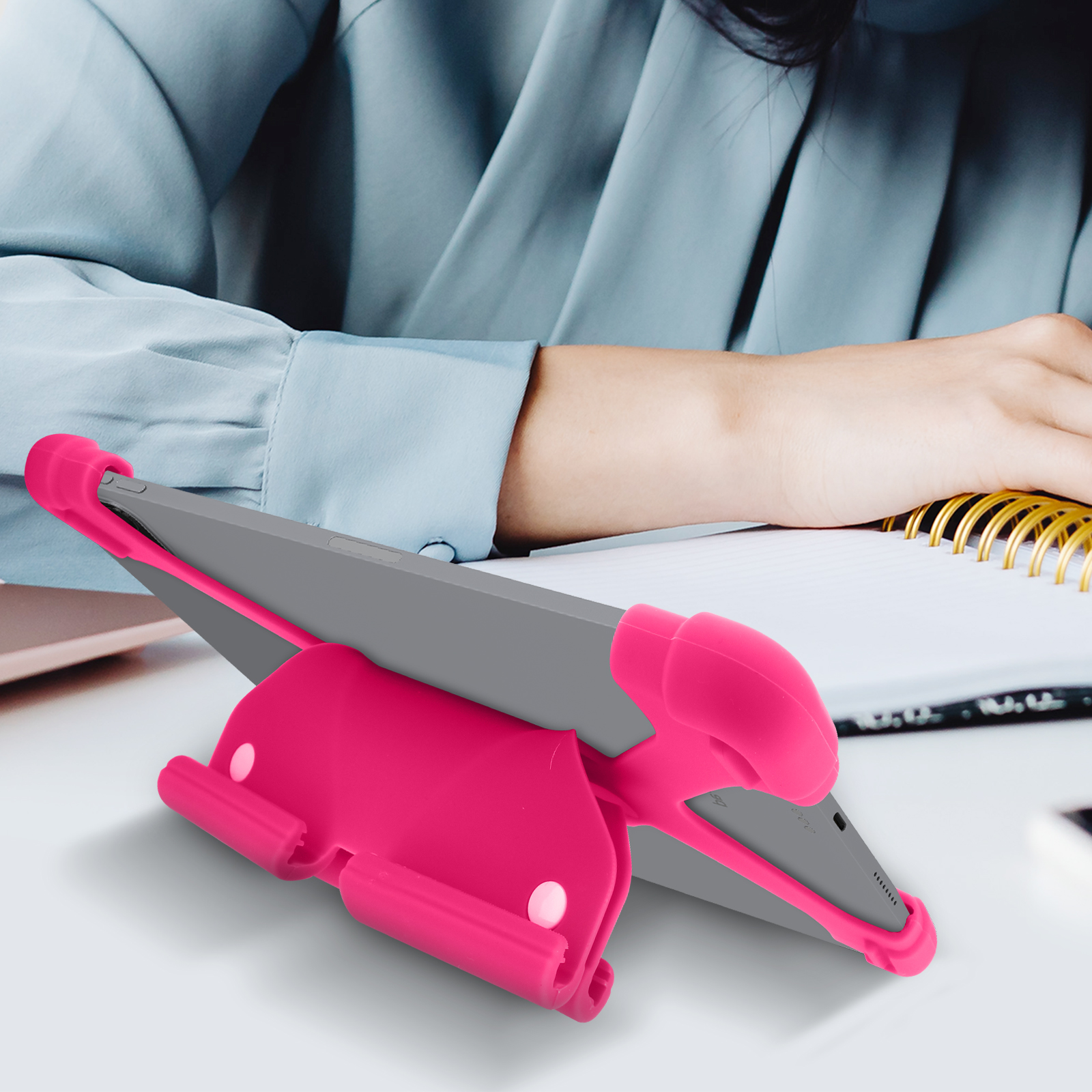 Un supporto per tablet rosa che supporta un tablet grigio, con le mani di una persona e un taccuino visibili.