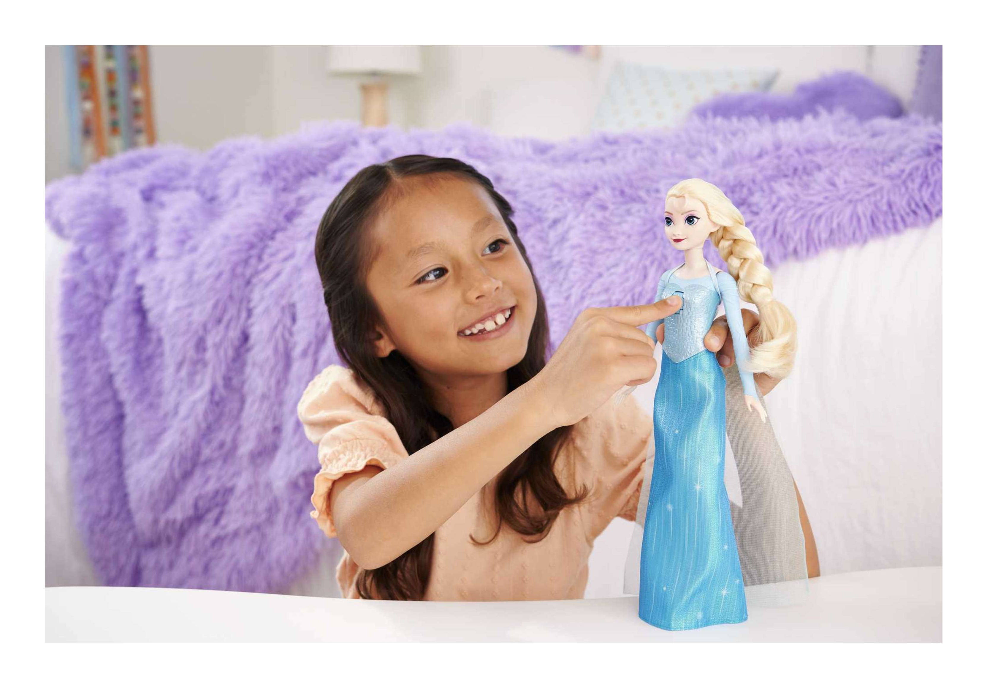 Mädchen zeigt auf eine Elsa-Puppe. Ein lila, flauschige Decke und ein Bett sind im Hintergrund.