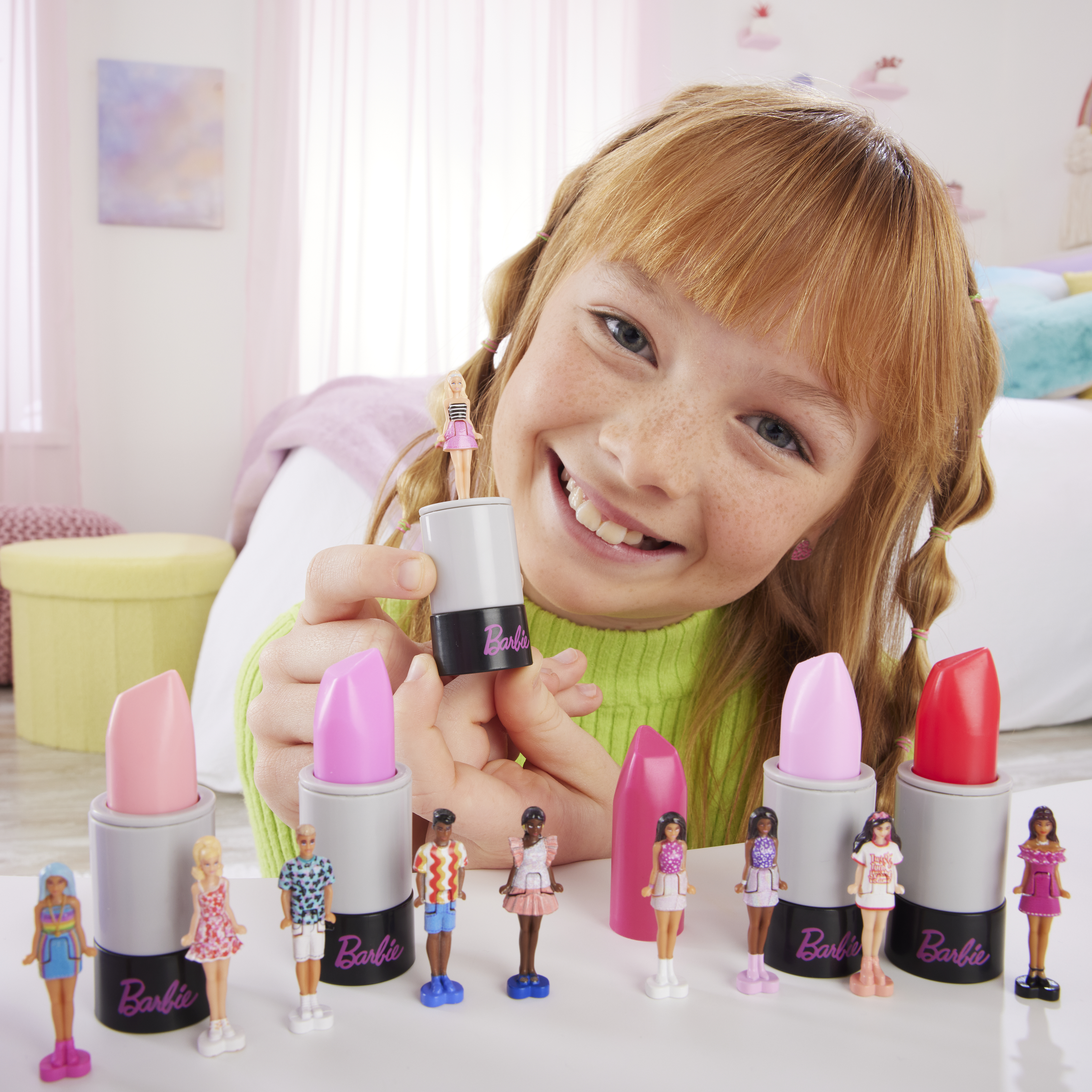 Mädchen hält Lippenstiftspielzeug. Mehrere Lippenstifte und Barbie-Puppen liegen auf dem Tisch. Das Mädchen lächelt.