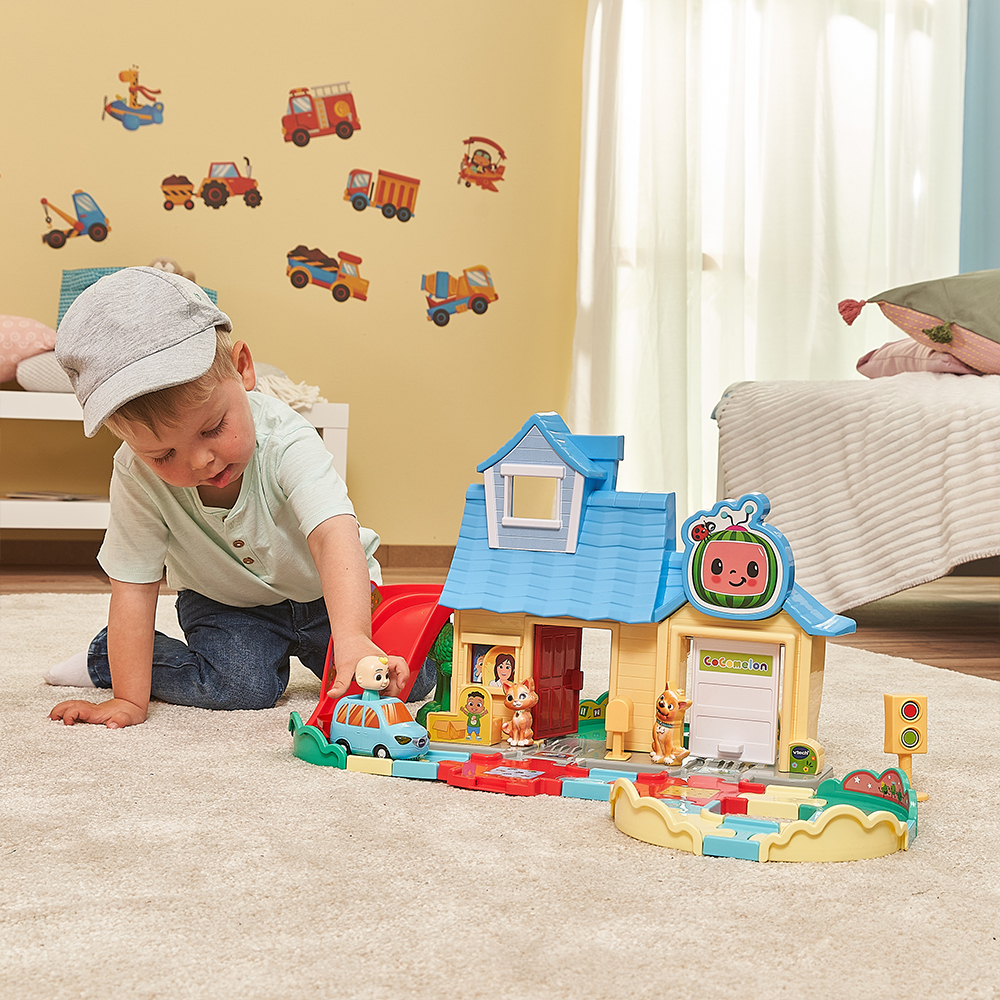 Ein Junge spielt mit einem Spielzeughaus und Autos. Das Haus hat eine Rutsche und steht auf einem Teppich mit Spielzeugautos.