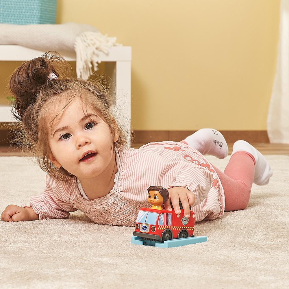 Mädchen spielt mit einem roten Spielzeug-Feuerwehrwagen auf einem Teppich, liegend.