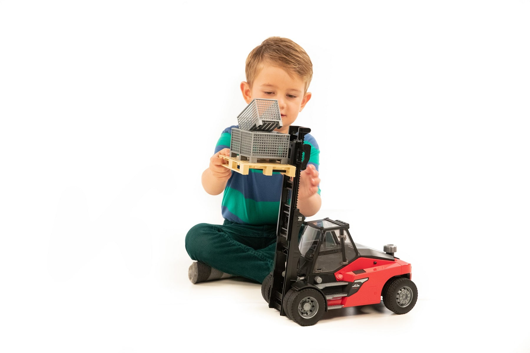 Dziecko bawi się zabawką wózkiem widłowym i paletami. Siedzi na białej podłodze.