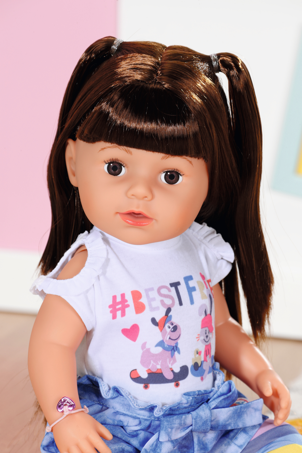 Eine Babypuppe mit braunen Zöpfen, trägt ein weißes T-Shirt und einen blauen Rock. Sie hat ein Armband.