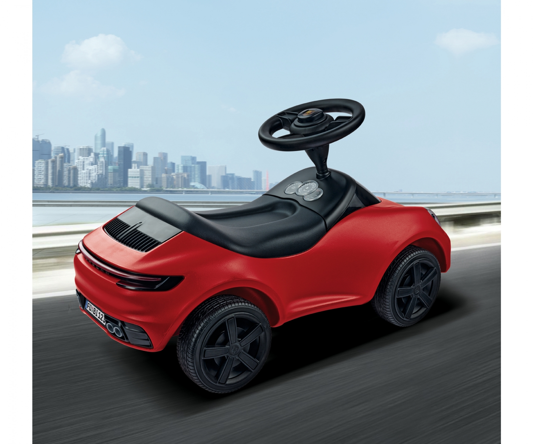 Rotes Spielzeugauto zum Aufsitzen mit schwarzen Rädern und Lenkrad, vor einer Stadt.