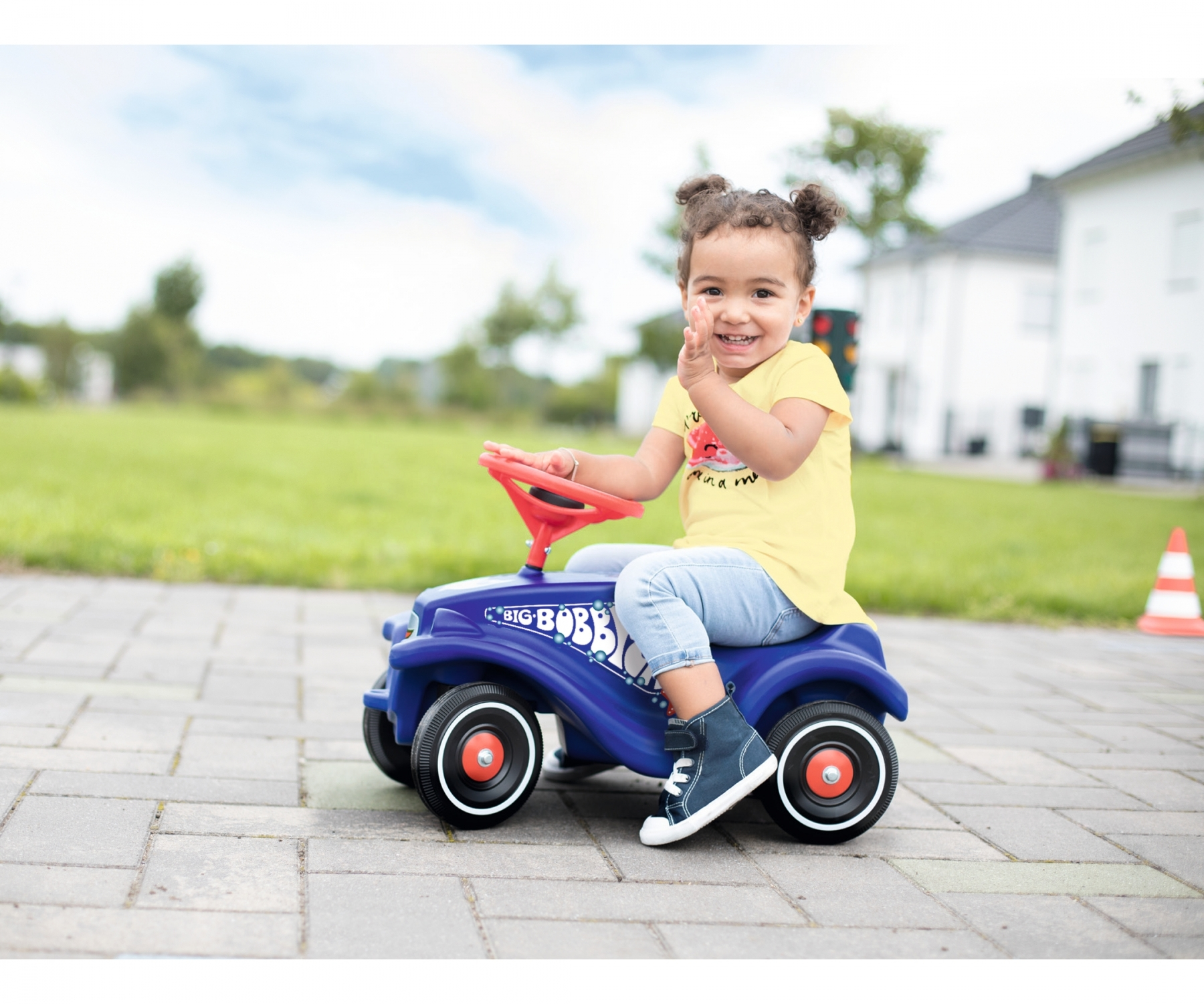 Ein junges Mädchen mit Zöpfen lächelt, während es auf einem blauen Spielzeugauto sitzt. Sie trägt ein gelbes T-Shirt.