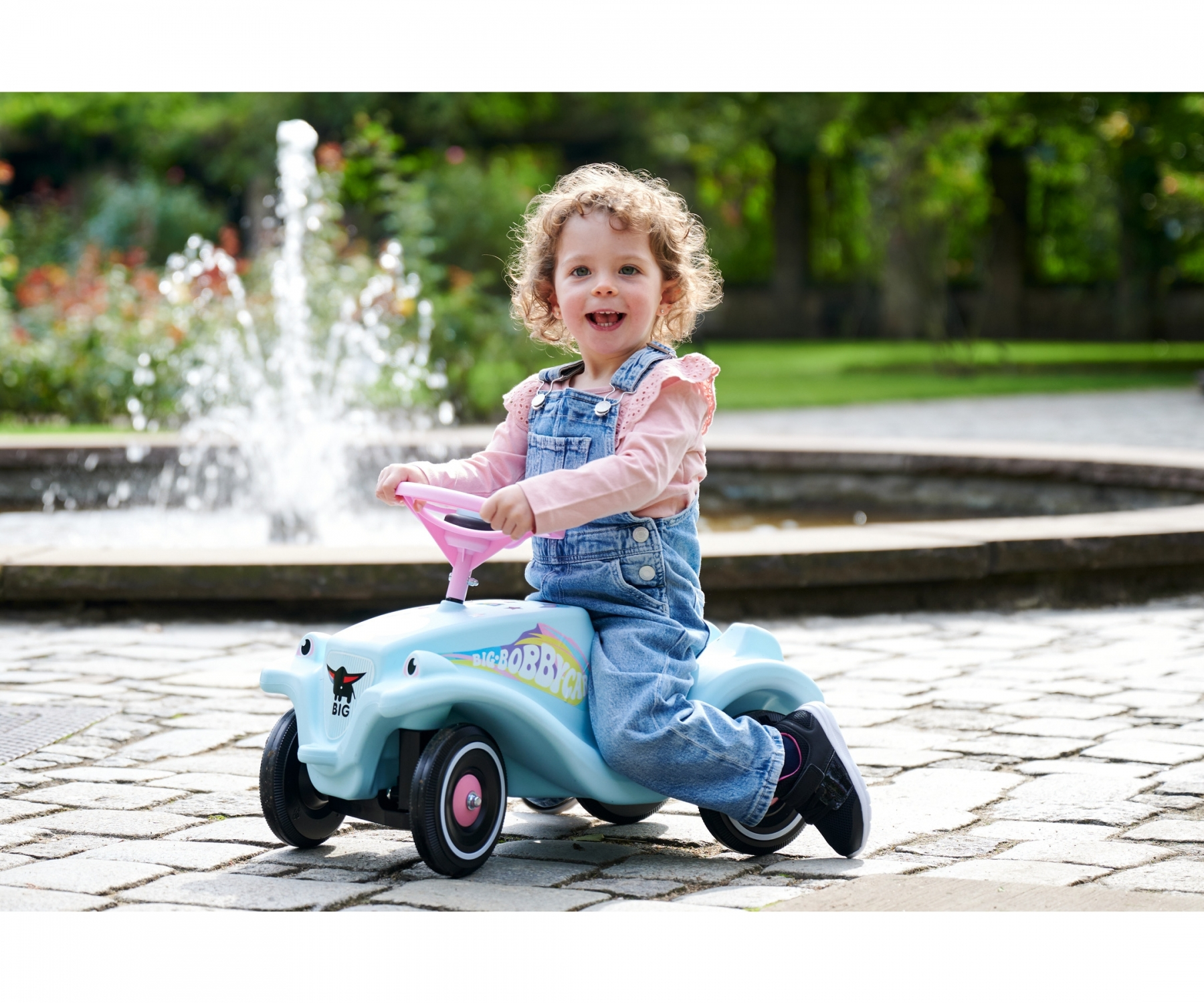 Una bambina su un'auto giocattolo blu in un parco. Una fontana è sullo sfondo.