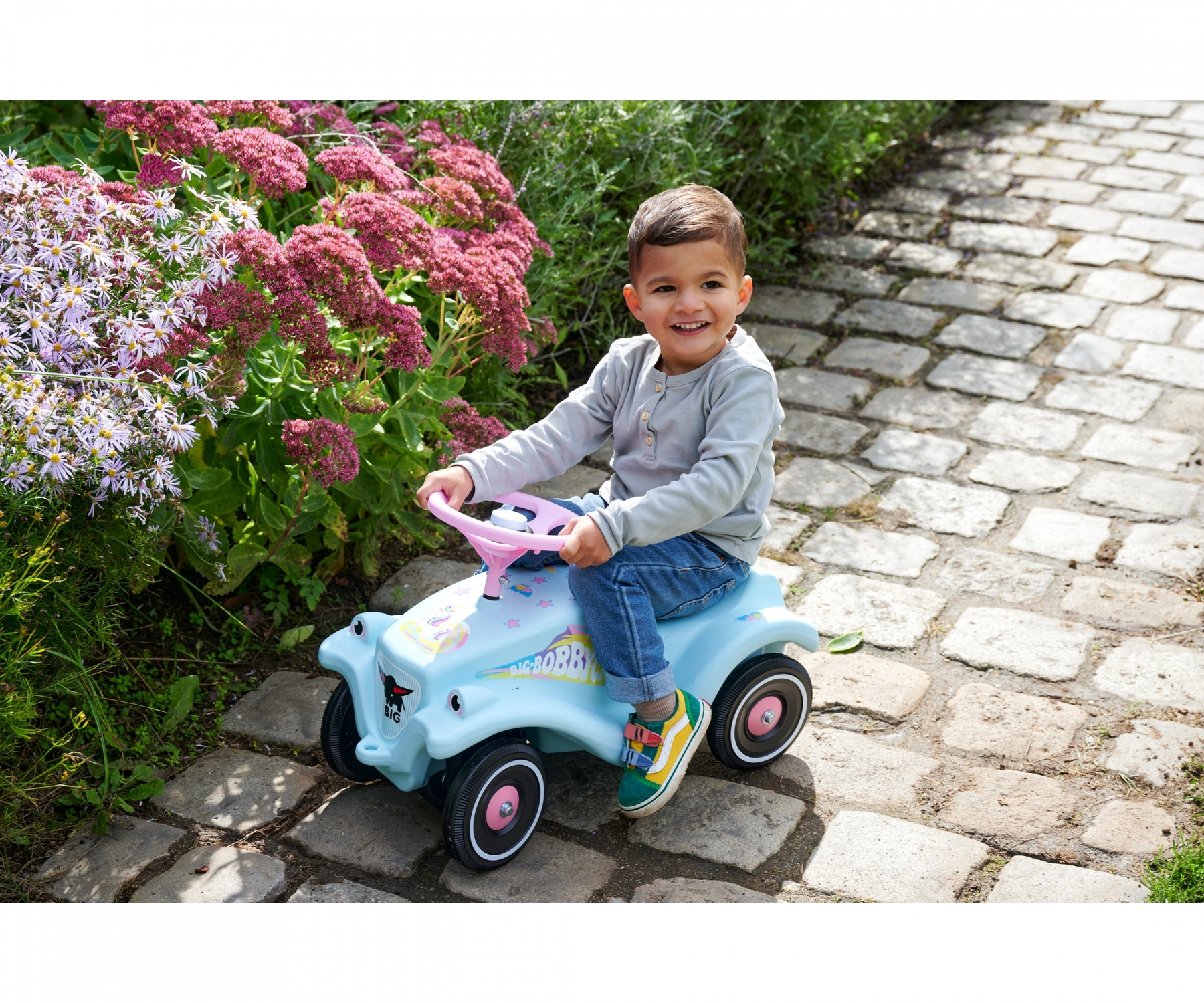 Ragazzo su un'auto giocattolo blu sorride, seduto su un sentiero di pietra, fiori sullo sfondo.
