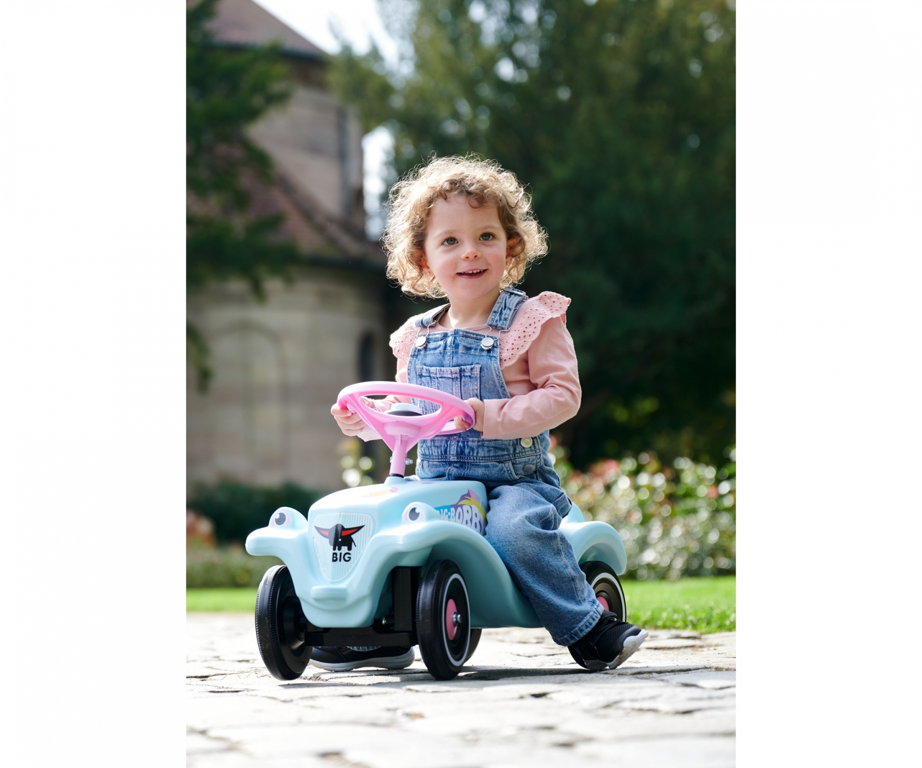 Un bambino piccolo guida un'auto giocattolo azzurra con un volante rosa. Una persona è sullo sfondo.