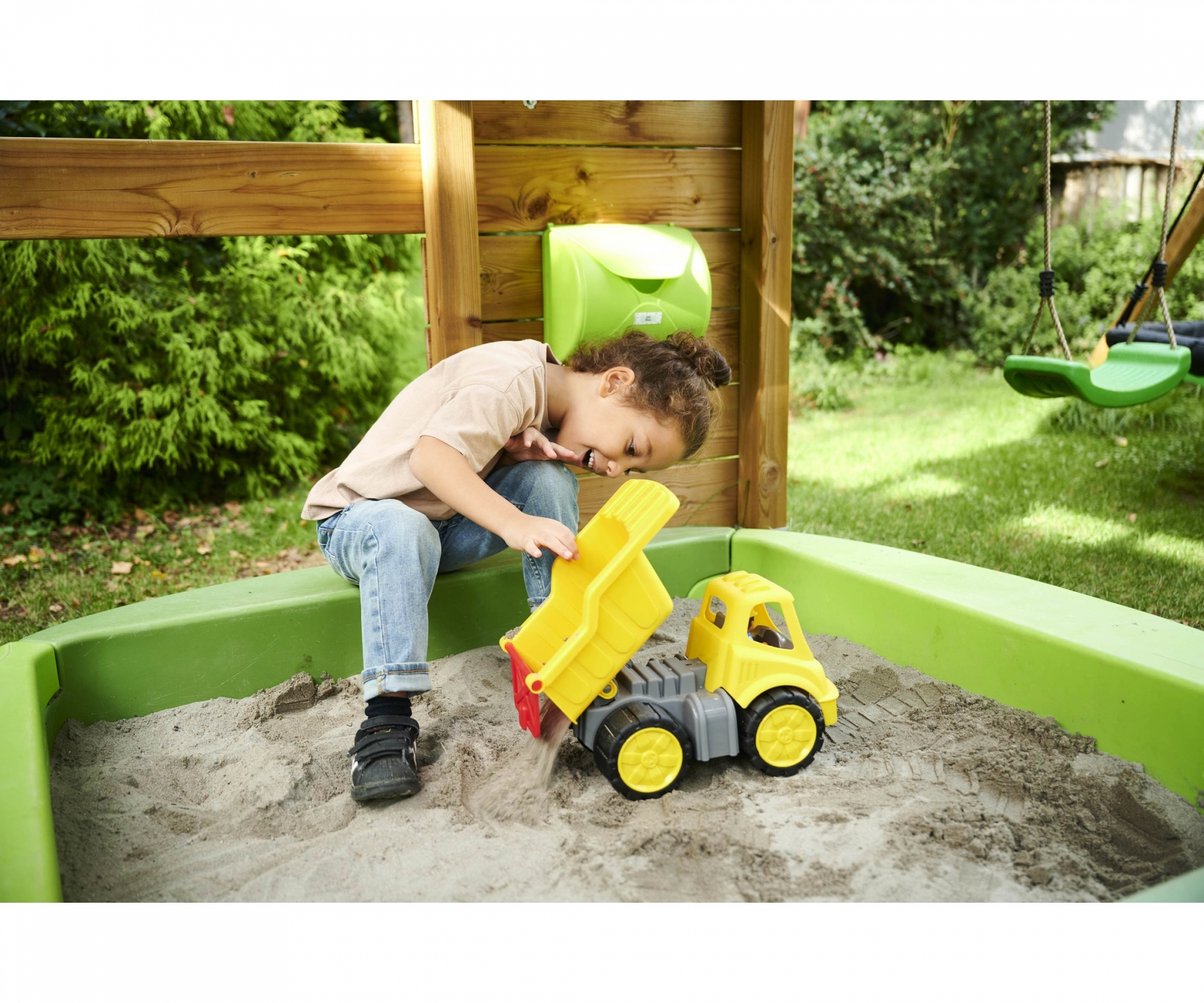 Kind spielt im Sandkasten mit einem gelben Spielzeug-LKW. Grüner Sandkasten, Holzstruktur im Hintergrund.