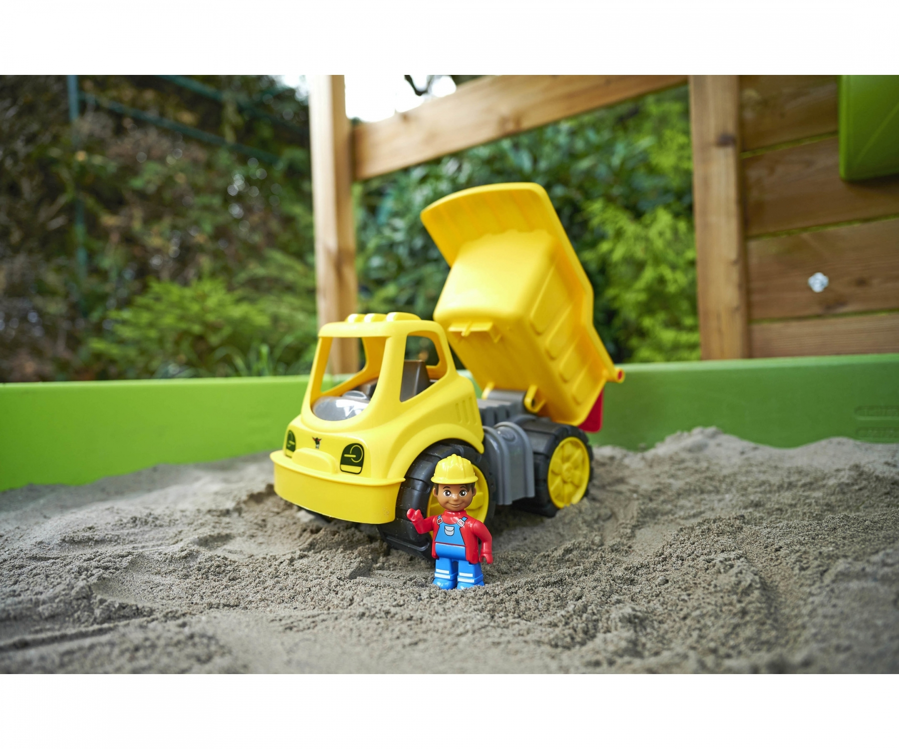 Ein gelber Spielzeug-Muldenkipper mit einer Bauarbeiter-Figur in einem Sandkasten.