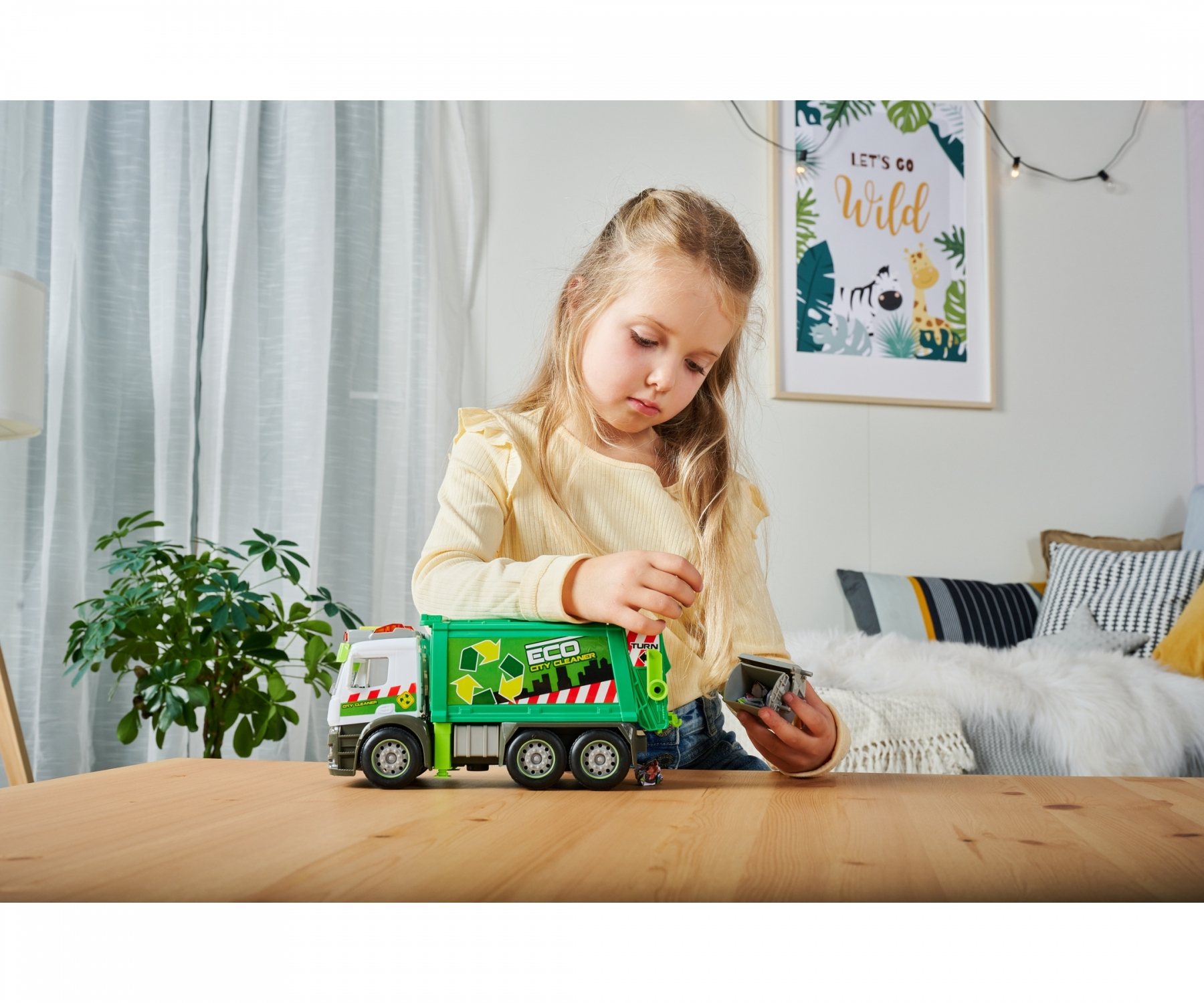 Mała dziewczynka bawi się zielono-czerwoną zabawką-śmieciarką na drewnianym stole.