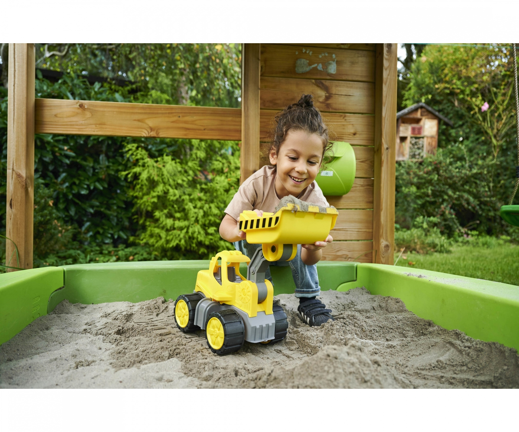 Dziecko bawi się żółtą zabawką-koparką w piaskownicy, uśmiechając się. W tle drewniana konstrukcja.