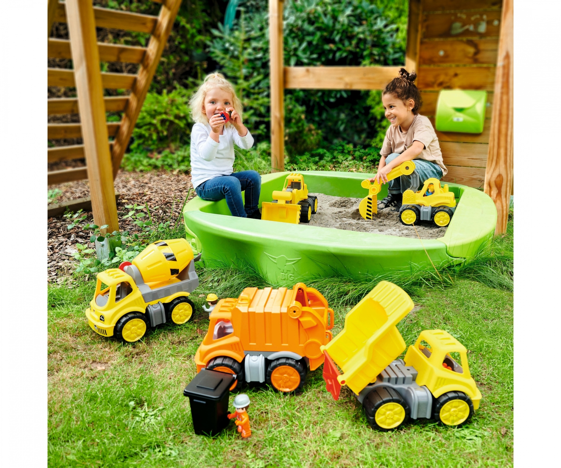 Dzieci bawią się zabawkowymi ciężarówkami w zielonej piaskownicy z drewnianą konstrukcją w tle.