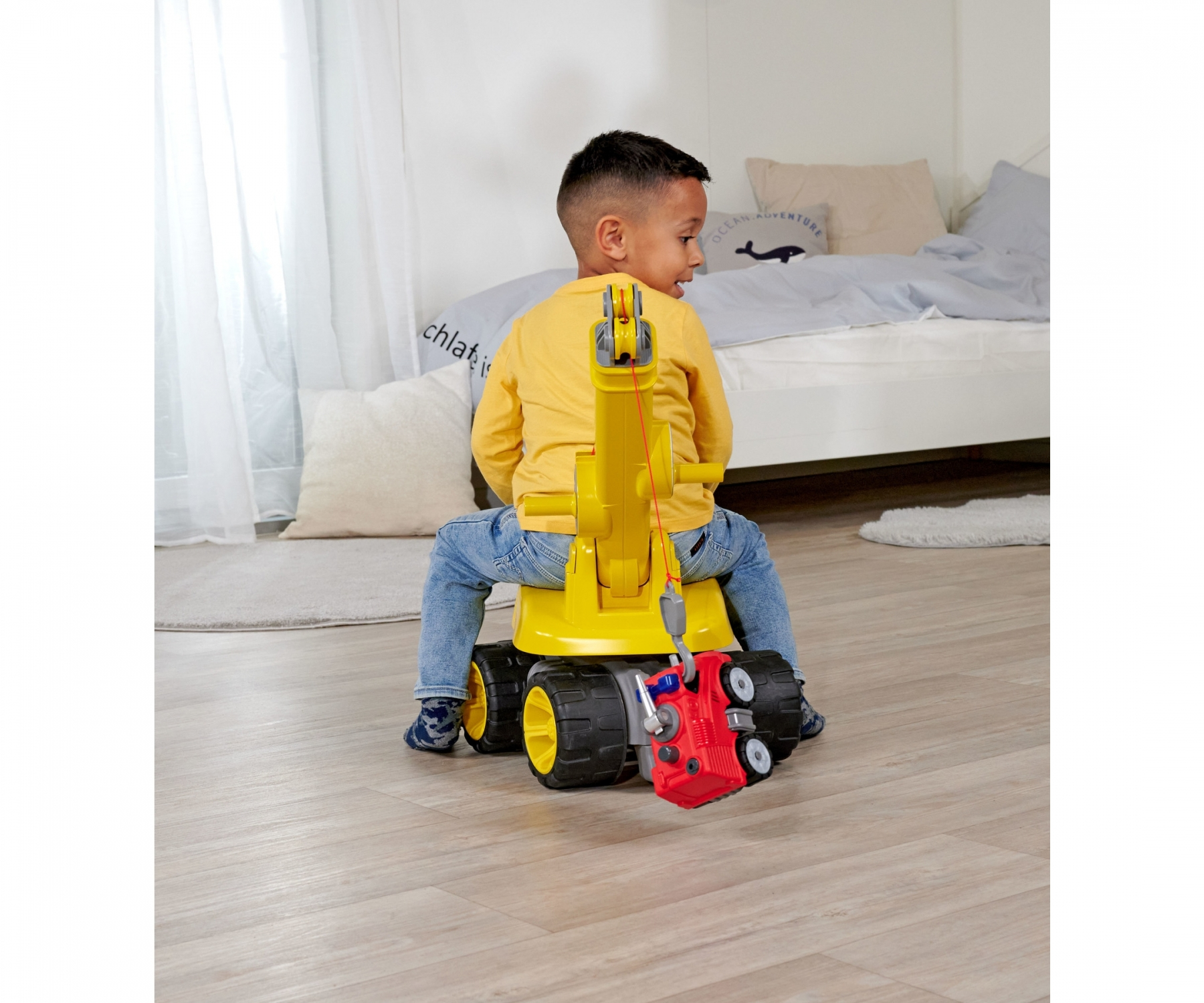 Dziecko siedzi na żółtym dźwigu-zabawce. Czerwony samochód jest zaczepiony do dźwigu.