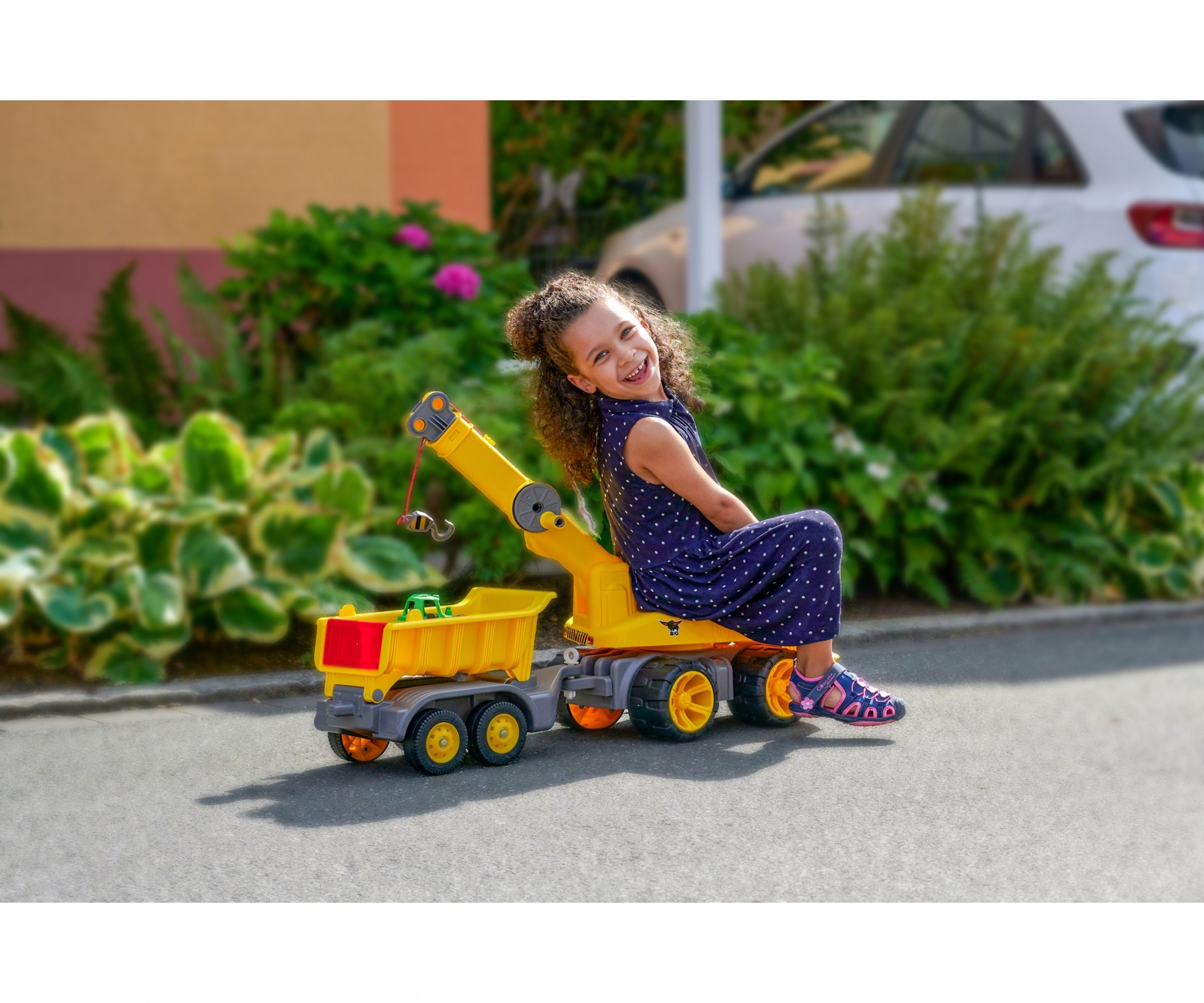 Uśmiechnięta dziewczynka siedząca na żółtej zabawce-ciężarówce z czerwoną przyczepą na drodze.