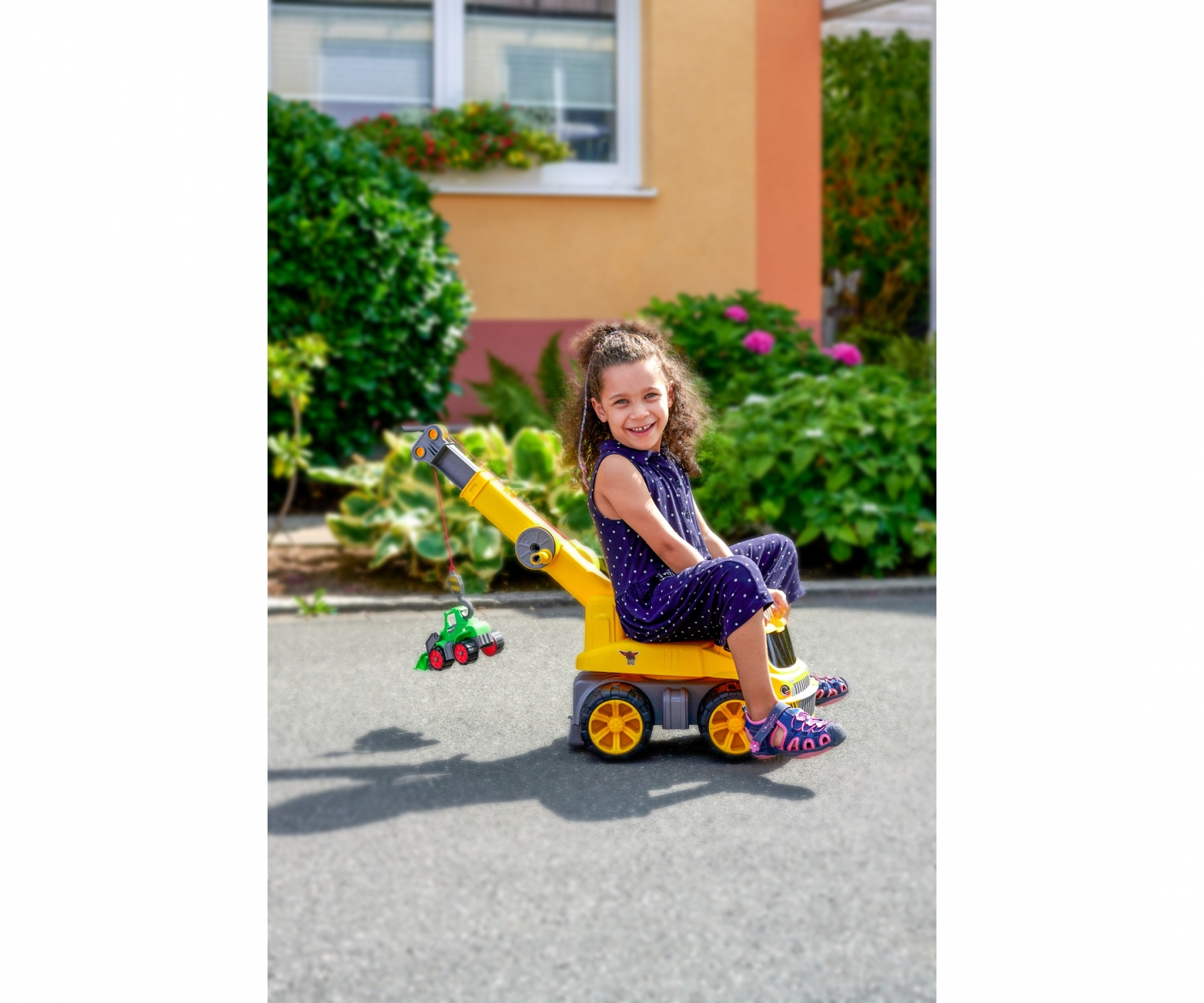 Dziewczynka na żółtym dźwigu. Zielony zabawkowy samochód wisi. Budynek i zielone krzewy w tle.