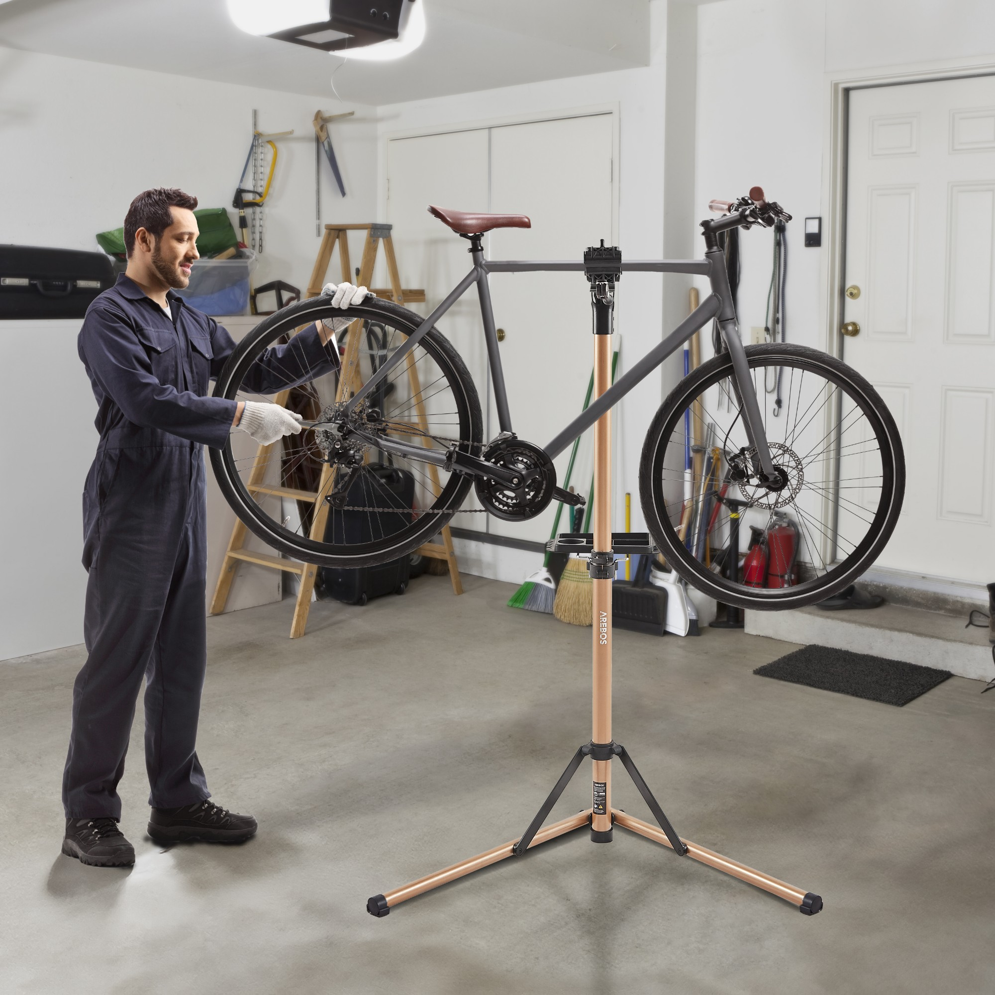 Mann in Overalls repariert graues Fahrrad auf Ständer in Garage. Hintergrund: Tür und Treppe.