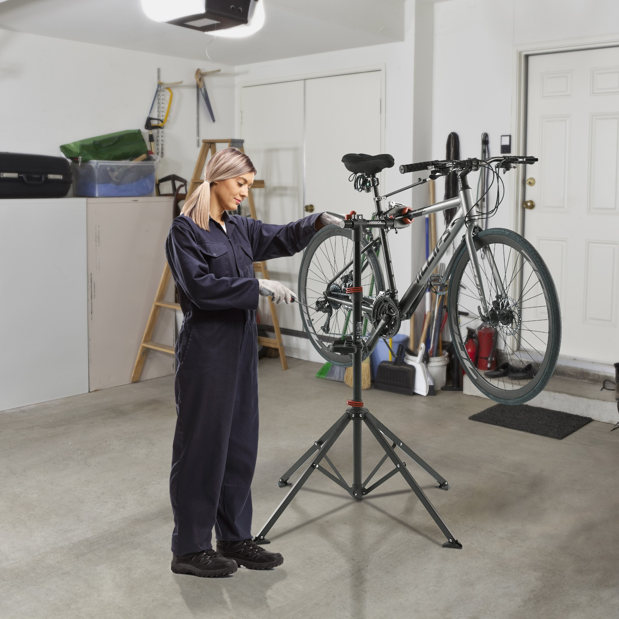Eine Frau in einem blauen Overall arbeitet in einer Garage an einem Fahrrad, das auf einem Reparaturständer montiert ist.