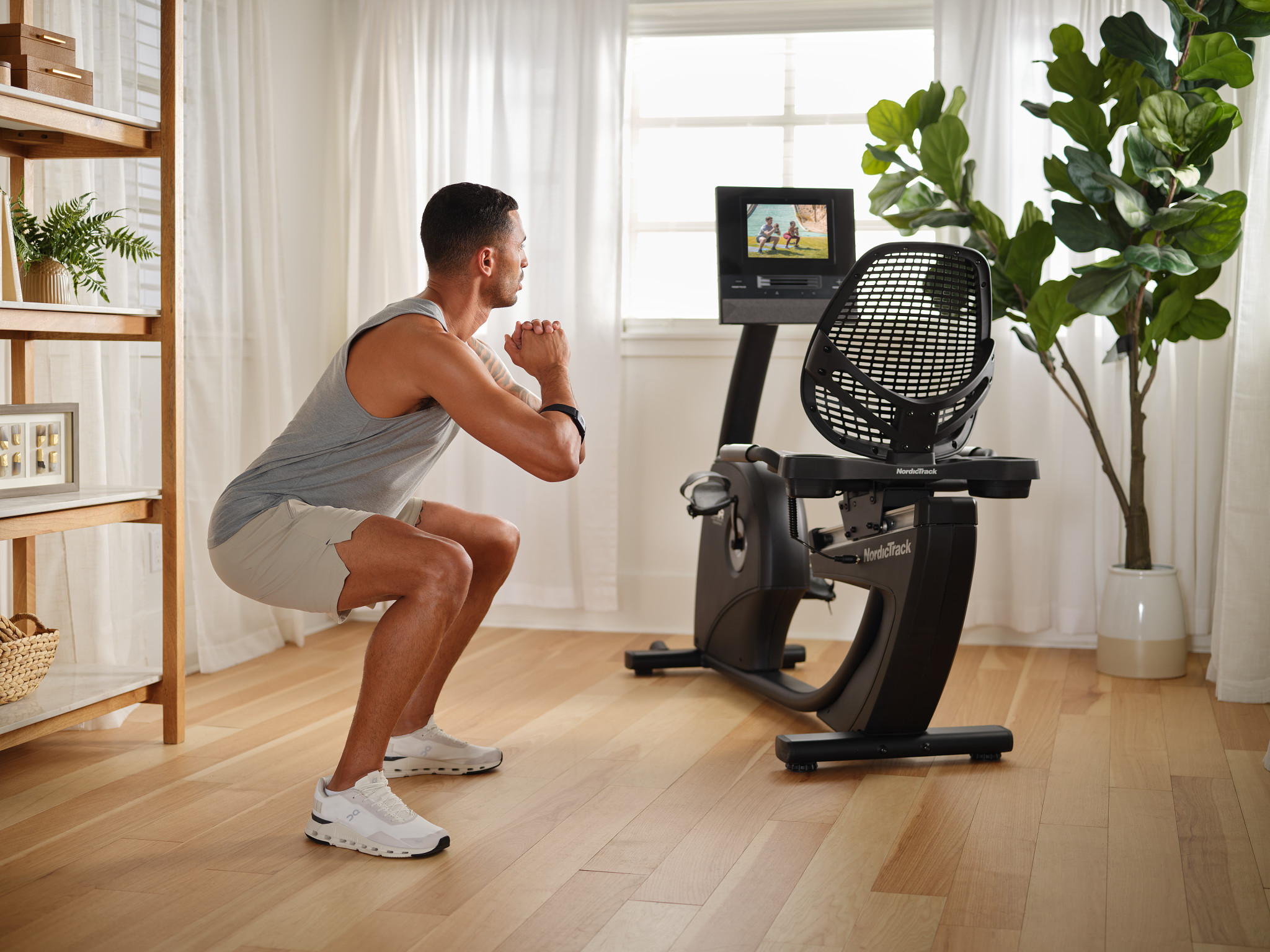 Ein Mann in einem grauen Tanktop und Shorts macht Kniebeugen vor einem Heimtrainer in einem hellen Raum.