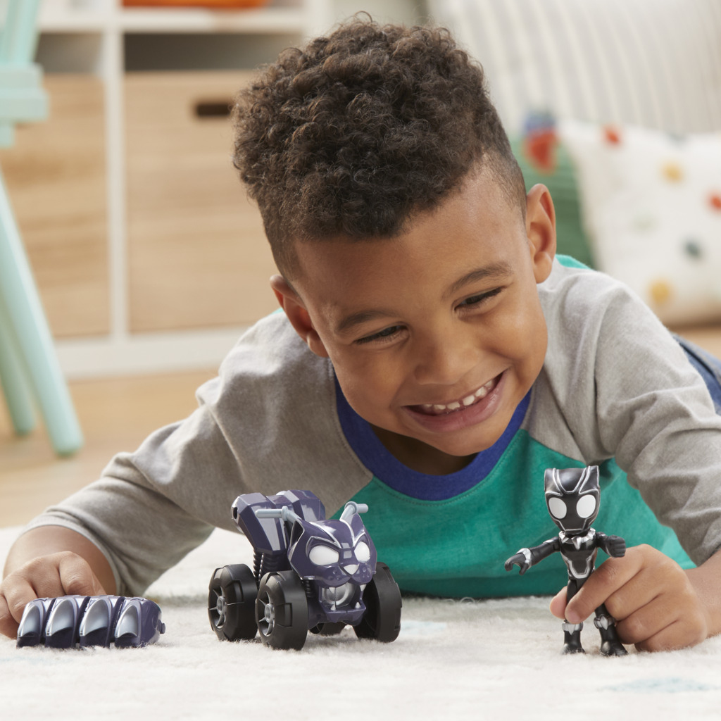 Chłopiec uśmiecha się, bawiąc się zabawkami Czarnej Pantery: samochodem i figurką. Dywan i meble w tle.