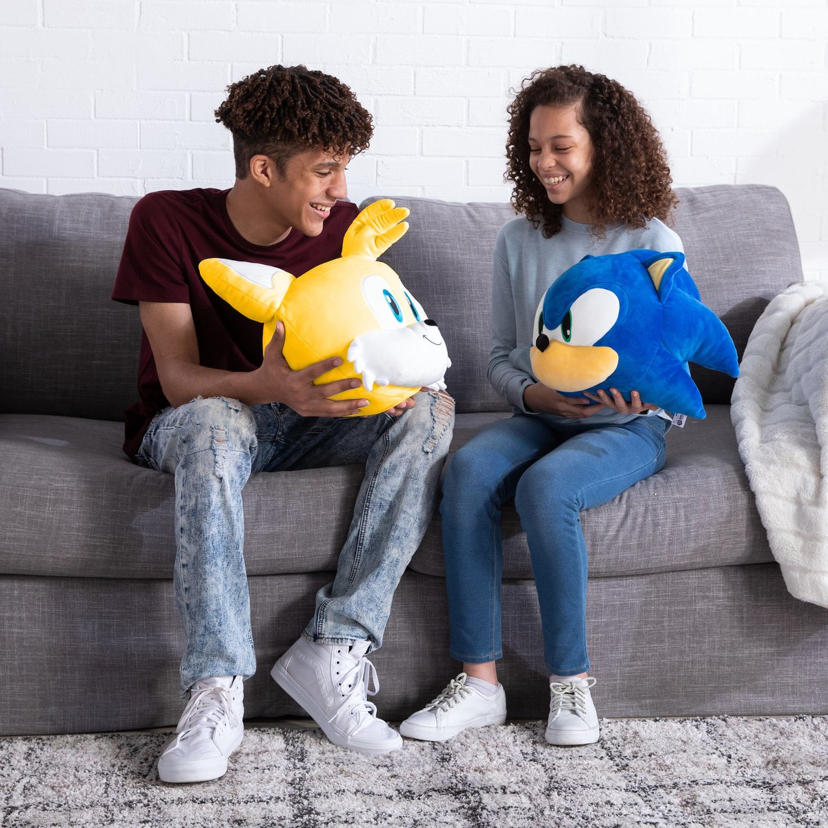 Due persone, ragazzo e ragazza, su divano grigio con peluche di Sonic e Tails, sorridono.