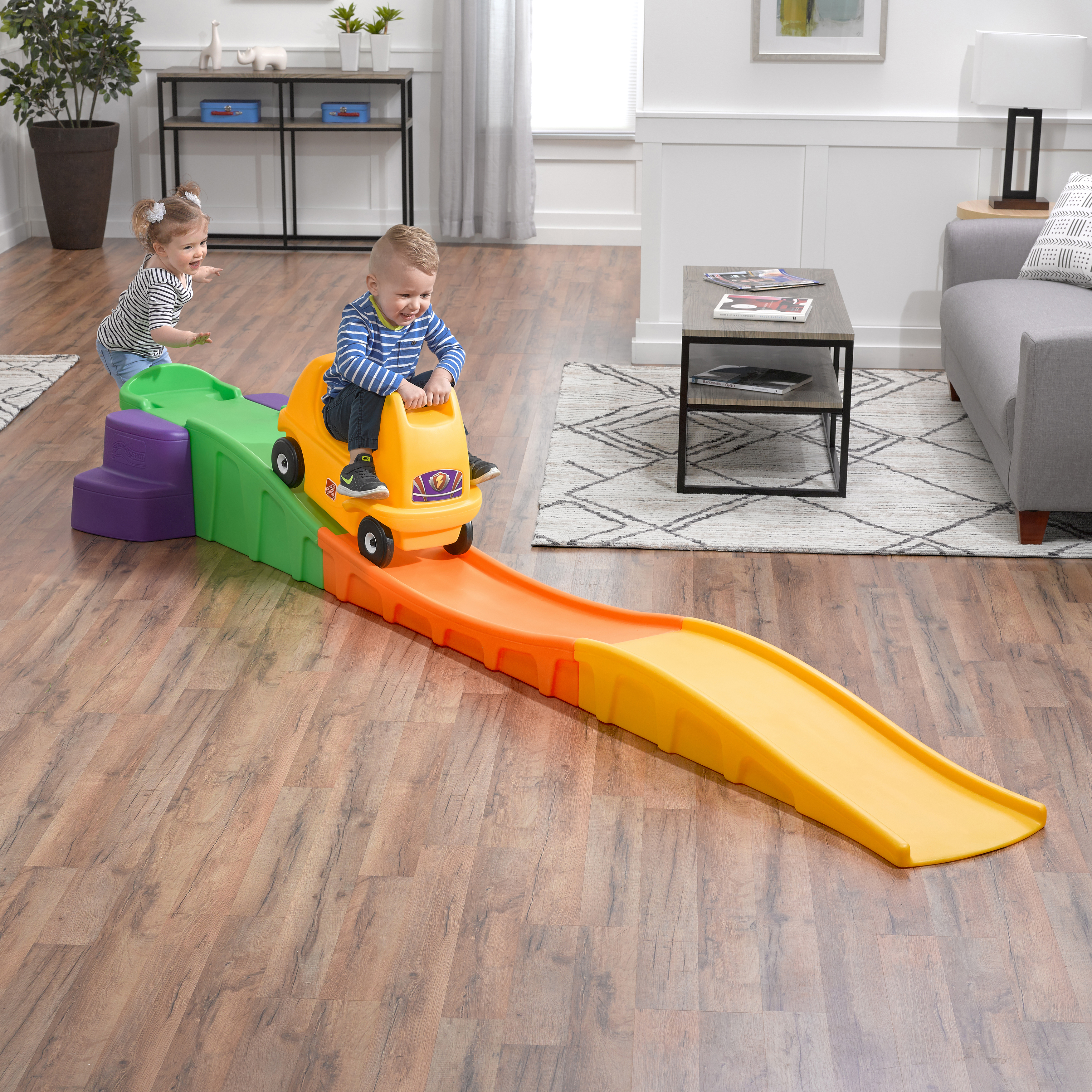 Zwei Kinder spielen drinnen auf einem Kunststoffauto und einer Rampenanlage, Holzboden, weiße Wand.
