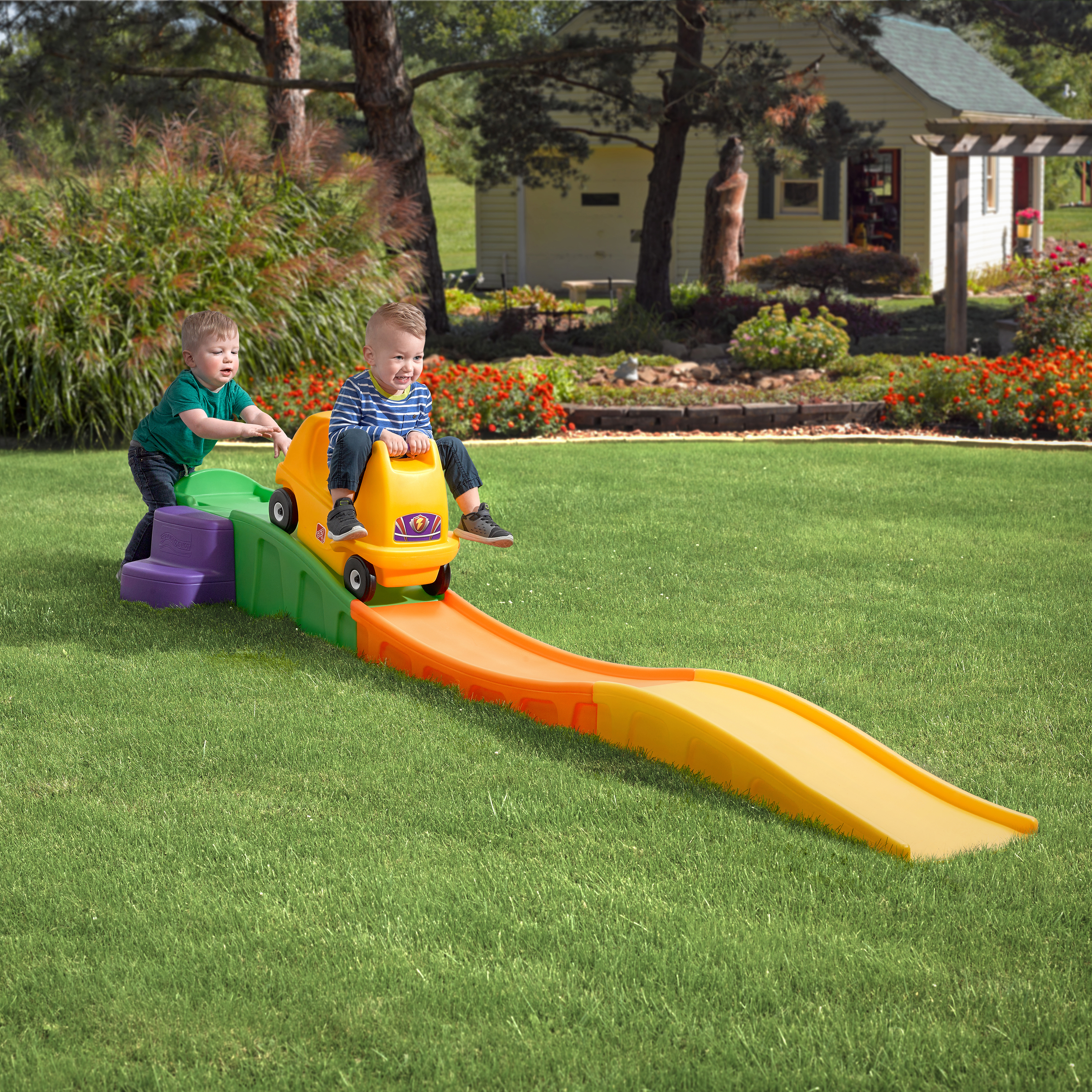 Zwei kleine Jungen spielen auf einer gelb-grünen Rutsche in einem grasbewachsenen Garten. Einer schiebt ein Spielzeugauto.