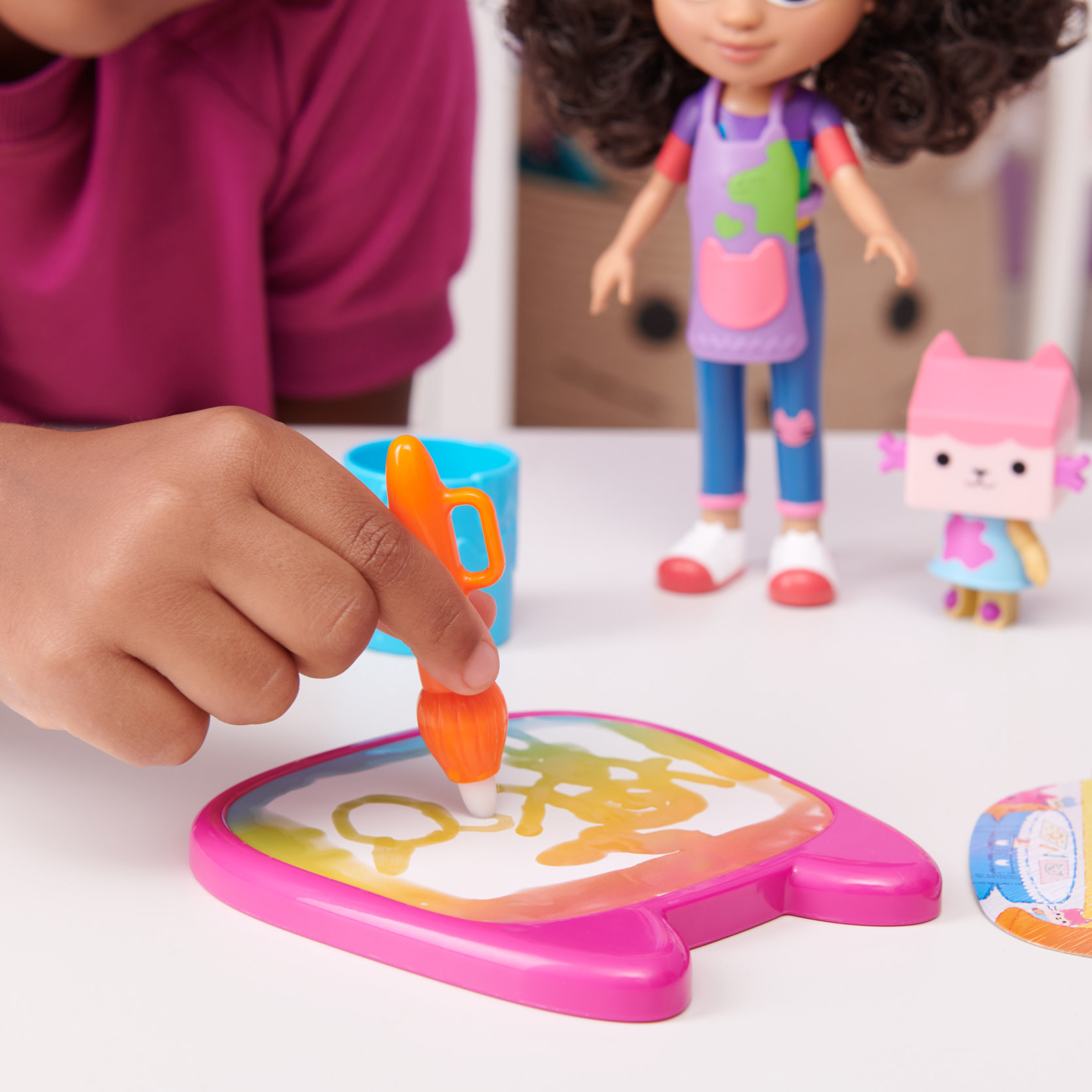 Dziecko rysuje pomarańczowym pędzlem na różowej tablicy. Jest lalka i zabawka dla kota.
