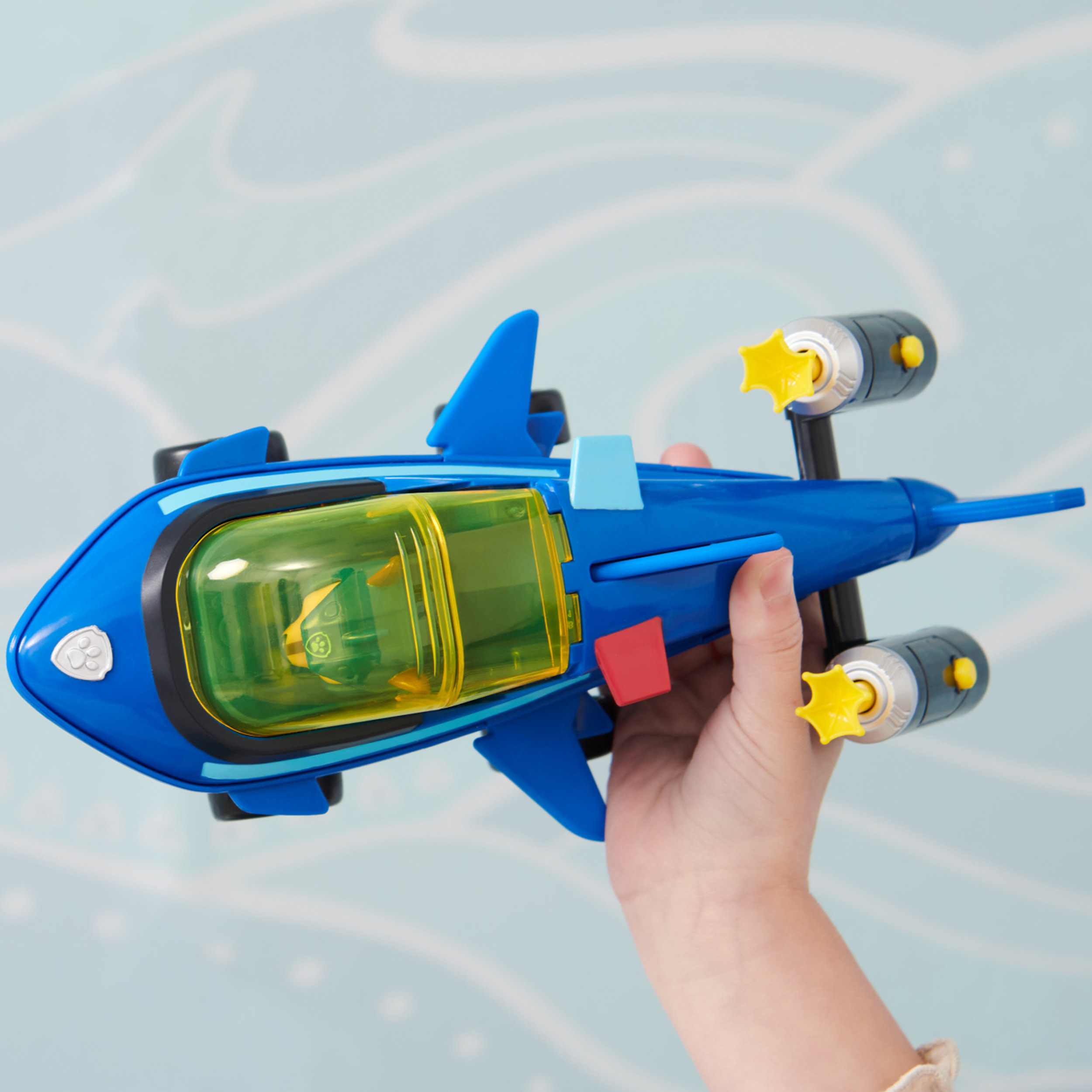 Blaues Spielzeugflugzeug mit gelben Akzenten, gehalten von einem Kind. Grünes und gelbes Fenster in der Mitte.