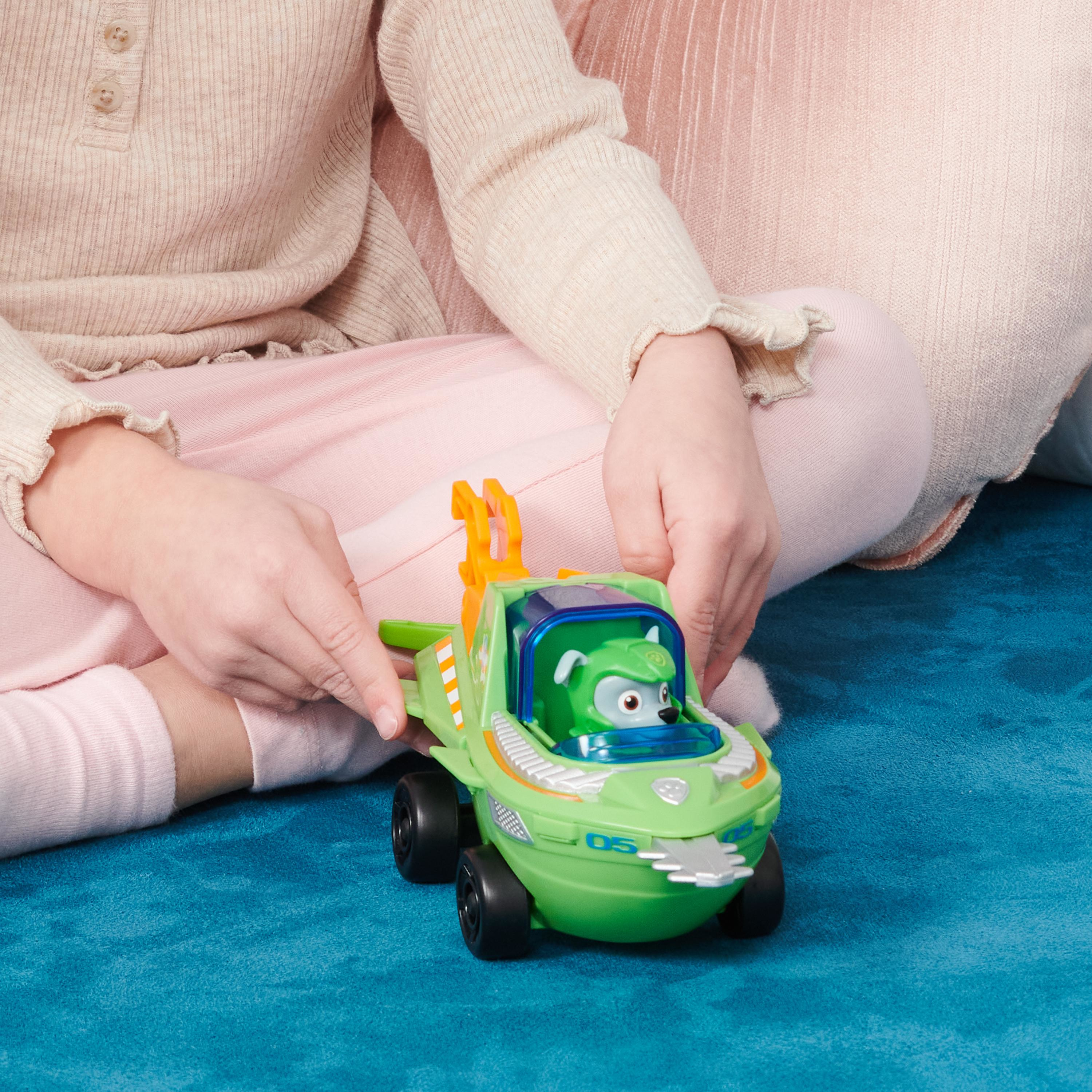 Kinderhände spielen mit einem grünen Spielzeugboot mit einer Zeichentrickfigur auf blauer Oberfläche.