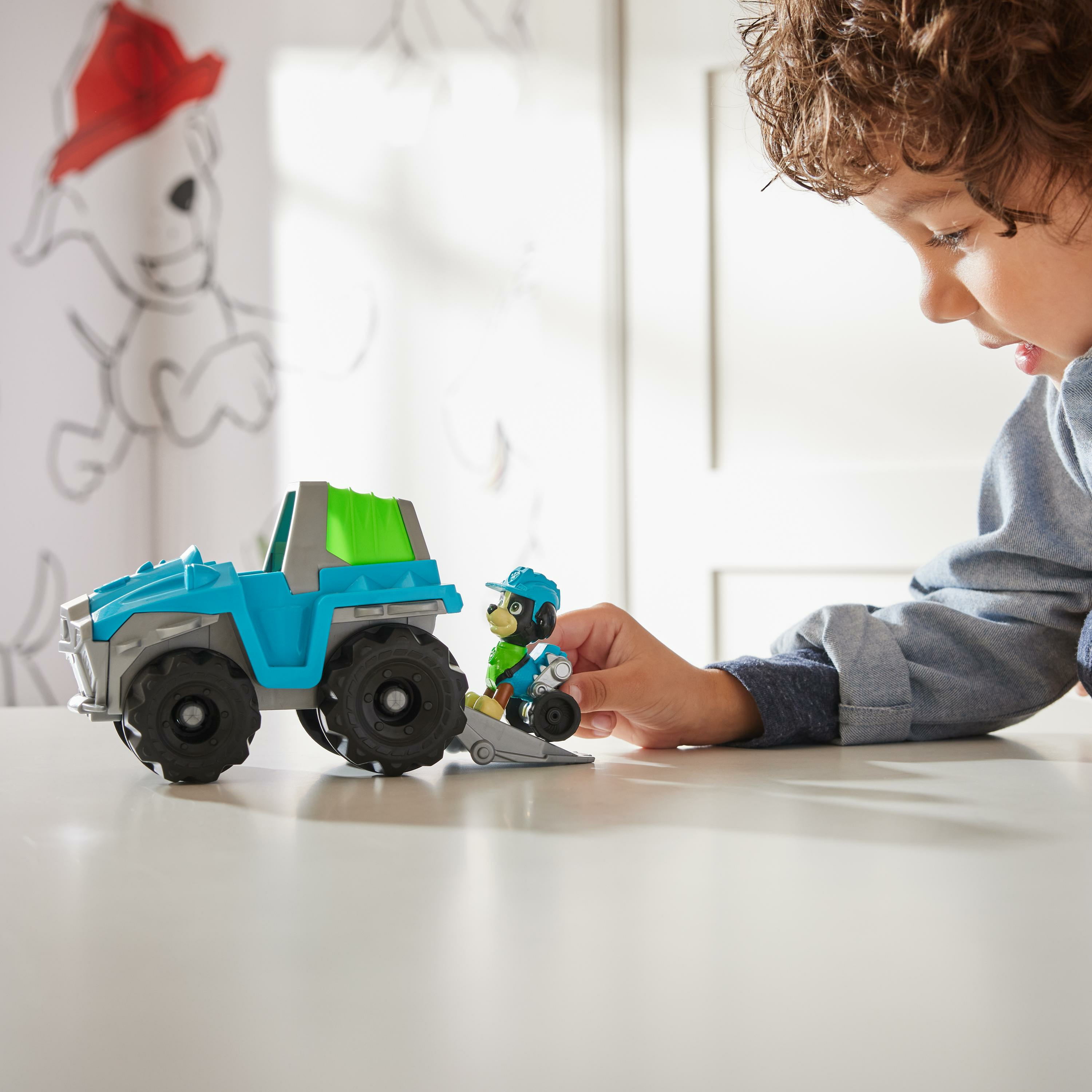 Ein Junge spielt mit einem blauen Spielzeug-LKW und einer kleinen Figur auf einem weißen Tisch.