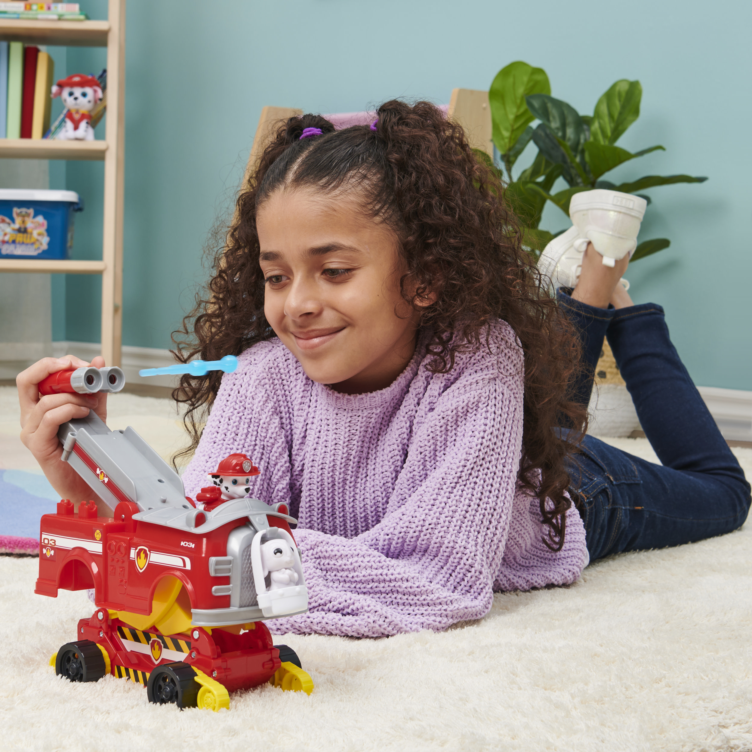 Ein Mädchen liegt auf einem Teppich und spielt mit einem Paw Patrol-Spielzeug. Ein Bücherregal ist im Hintergrund.