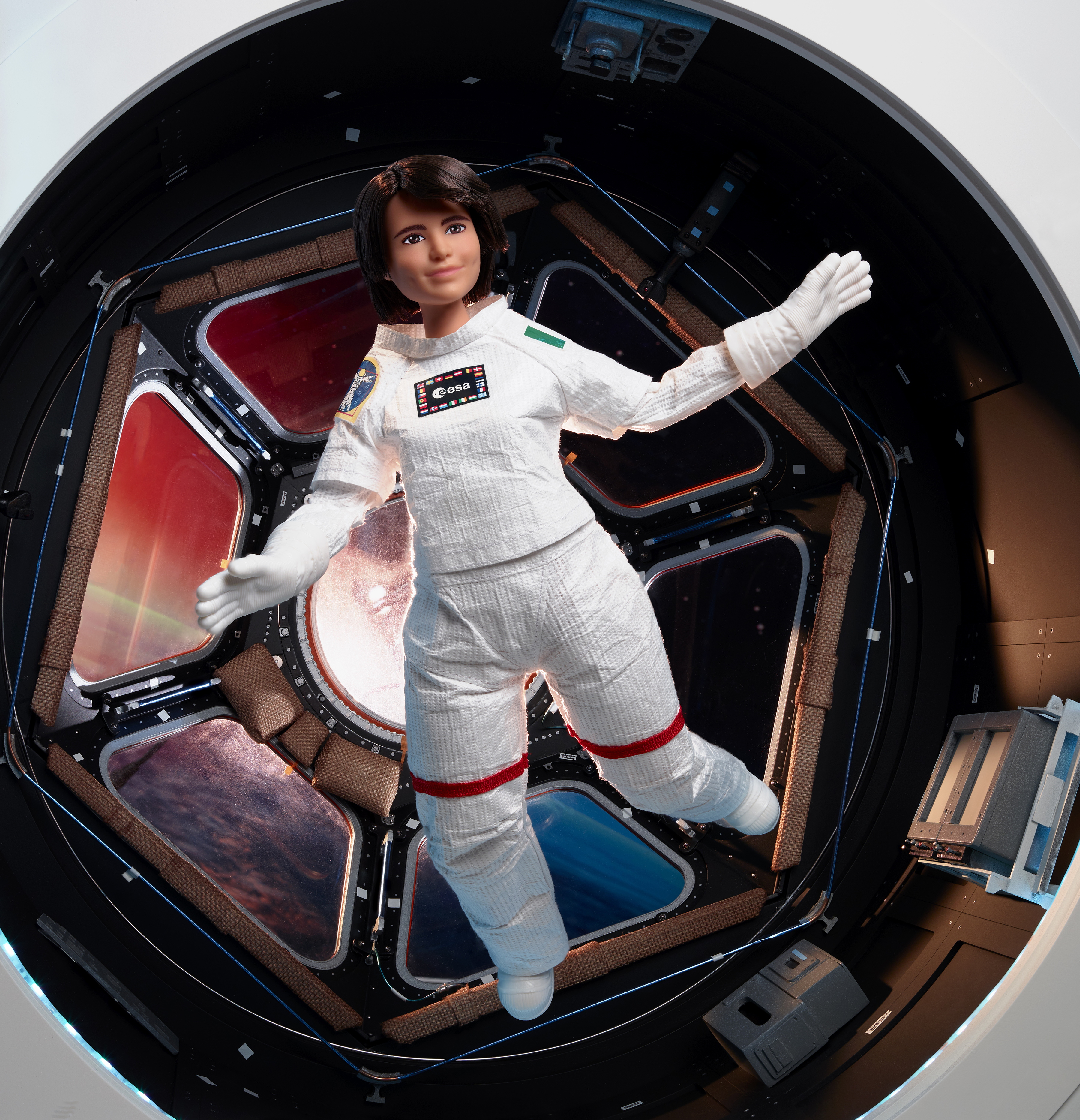 Una muñeca con traje espacial flota dentro de un modelo de estación espacial, con los brazos extendidos.