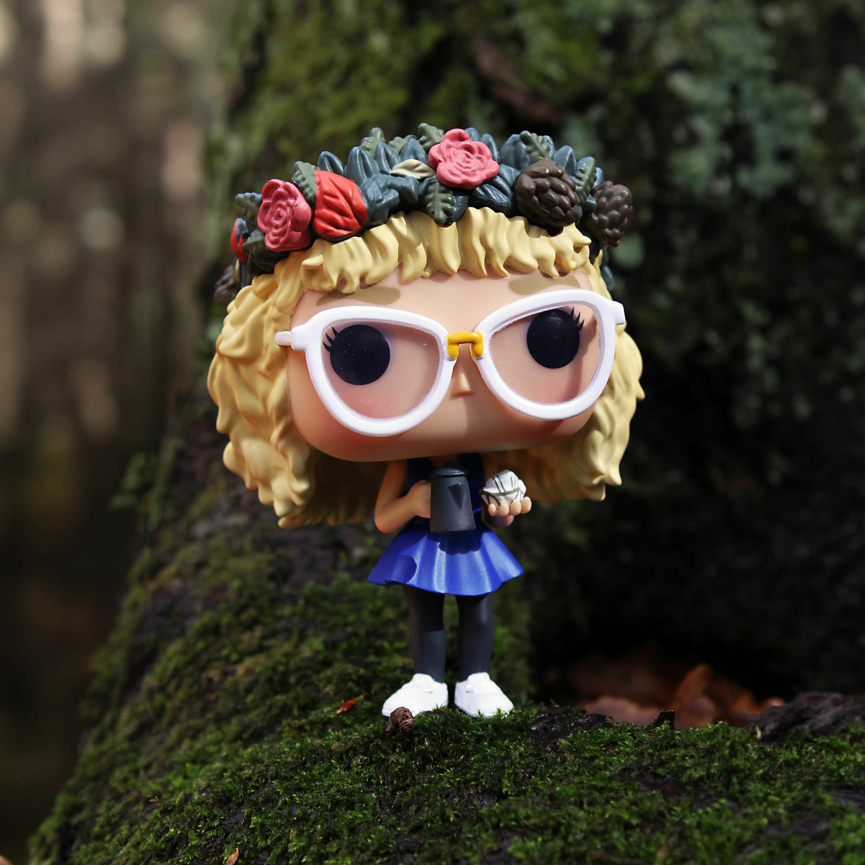 Funko Pop-Figur einer Frau mit Brille und Blumen. Im Hintergrund verschwommene grüne Bäume.